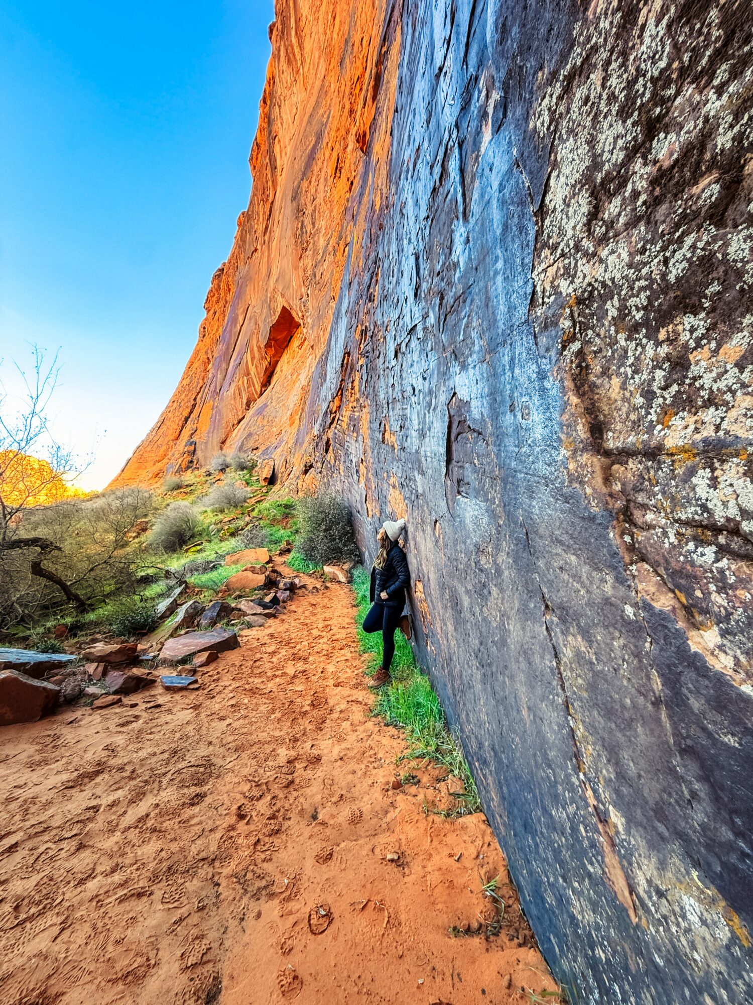 Person hiking along a trail beside a large rock face with a red cliff in the background, blue sky above.
