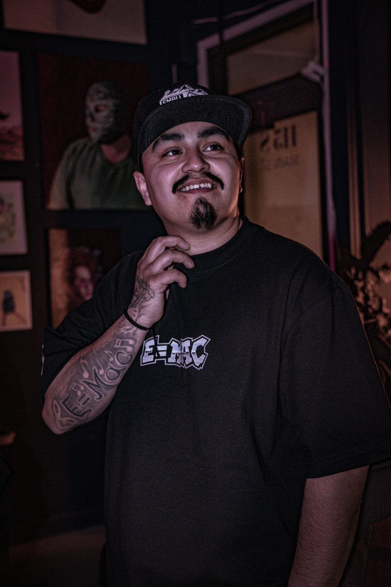 Man with a beard and tattooed arm wearing a black cap and T-shirt, smiling in a dimly lit room with portraits on the wall.