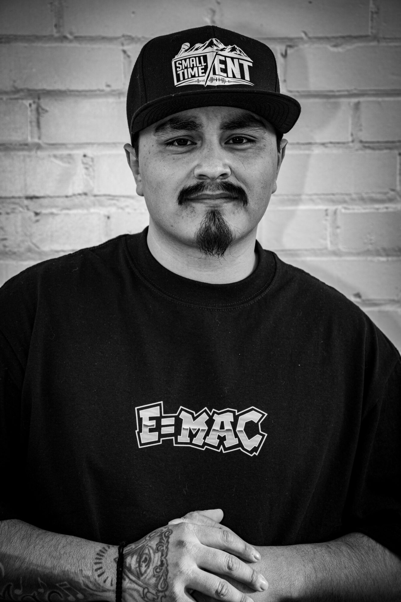 Man with a beard and mustache wearing a cap and T-shirt, standing against a brick wall.