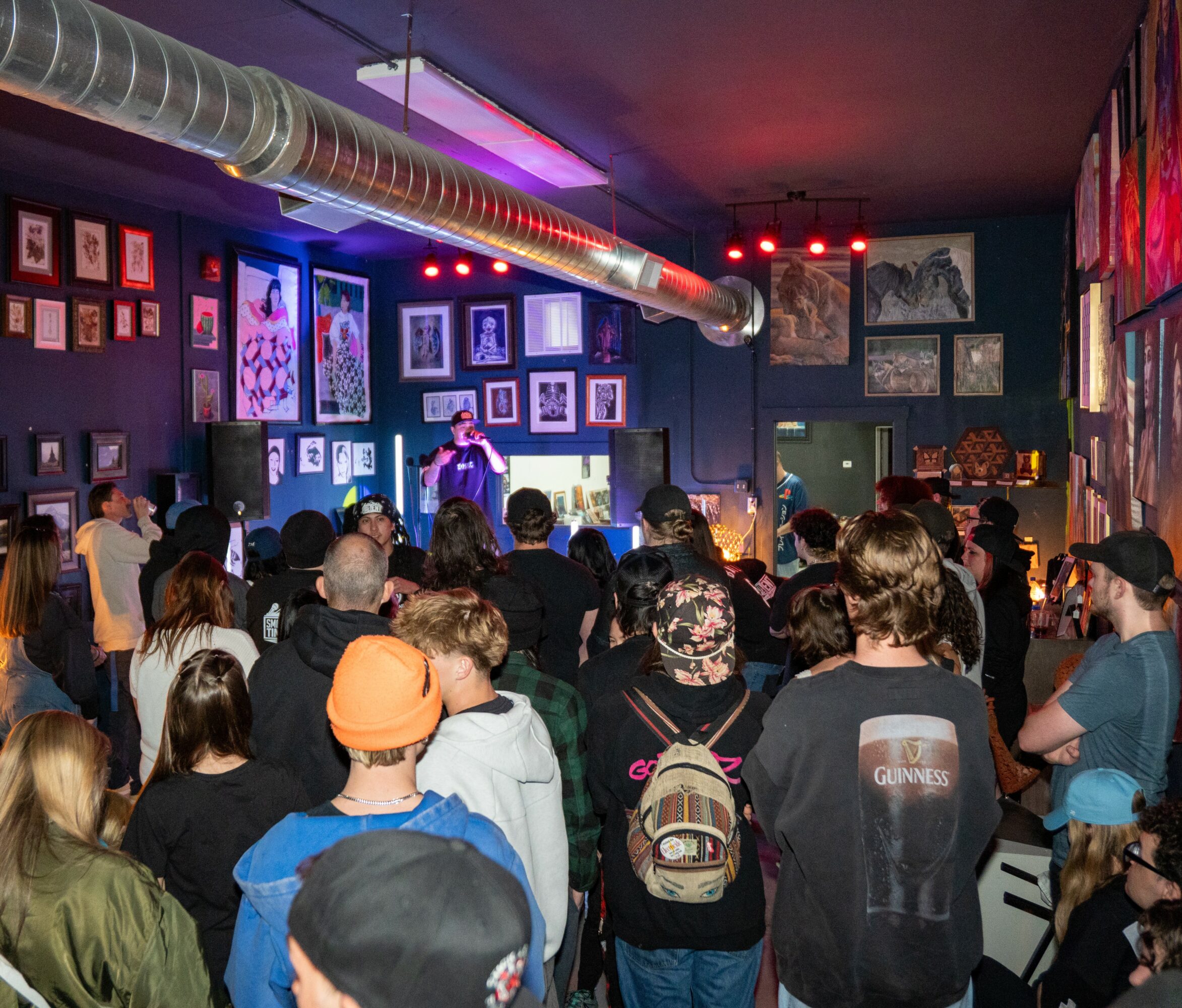 Crowd watching a performer on stage in an art gallery with framed artwork on dark walls, colorful lighting, and a ceiling vent.
