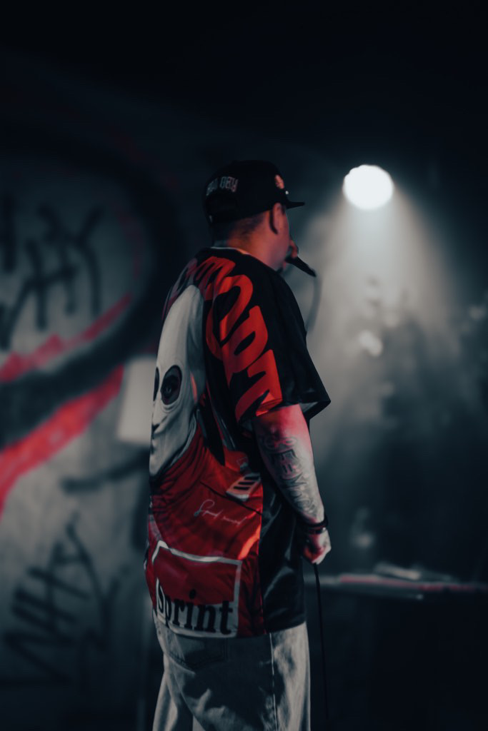 Person with tattoos and cap standing in a dark, graffiti-covered room illuminated by a bright light.