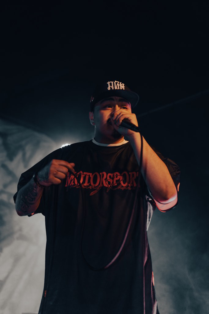 Person performing on stage with microphone, wearing a black cap and T-shirt, dark background, stage lighting, and smoke effects.