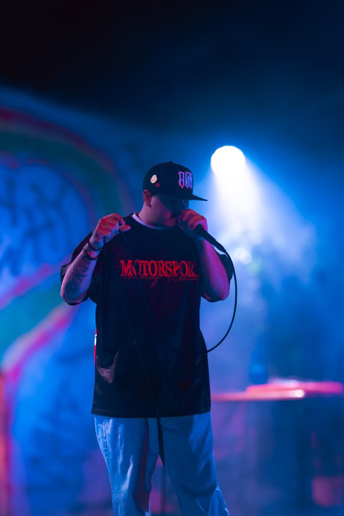 Person wearing a black cap and T-shirt holding a microphone on stage with blue and purple lighting