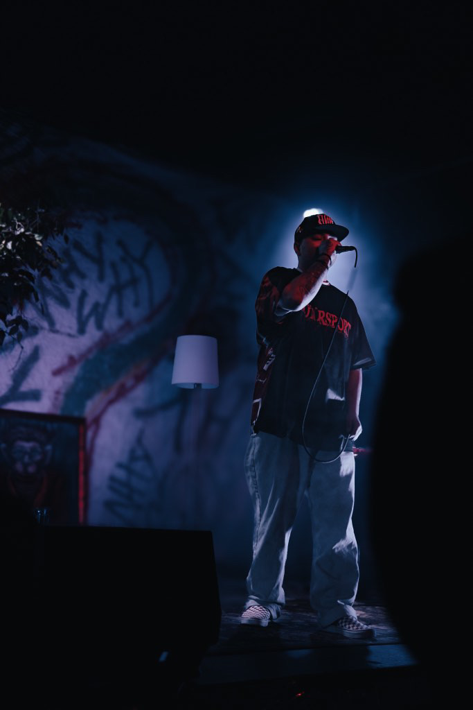 Person performing on stage with microphone, illuminated by blue light, graffiti background, wearing a cap and casual clothing.