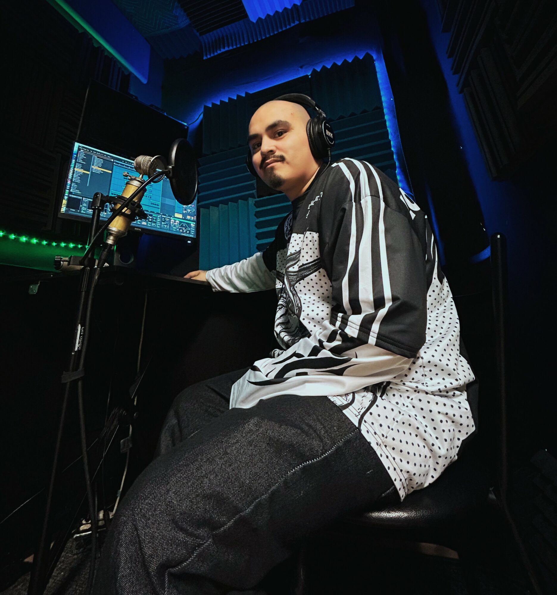 Person wearing headphones sitting in a recording studio with a microphone and computer screen, blue lighting, and soundproof walls.