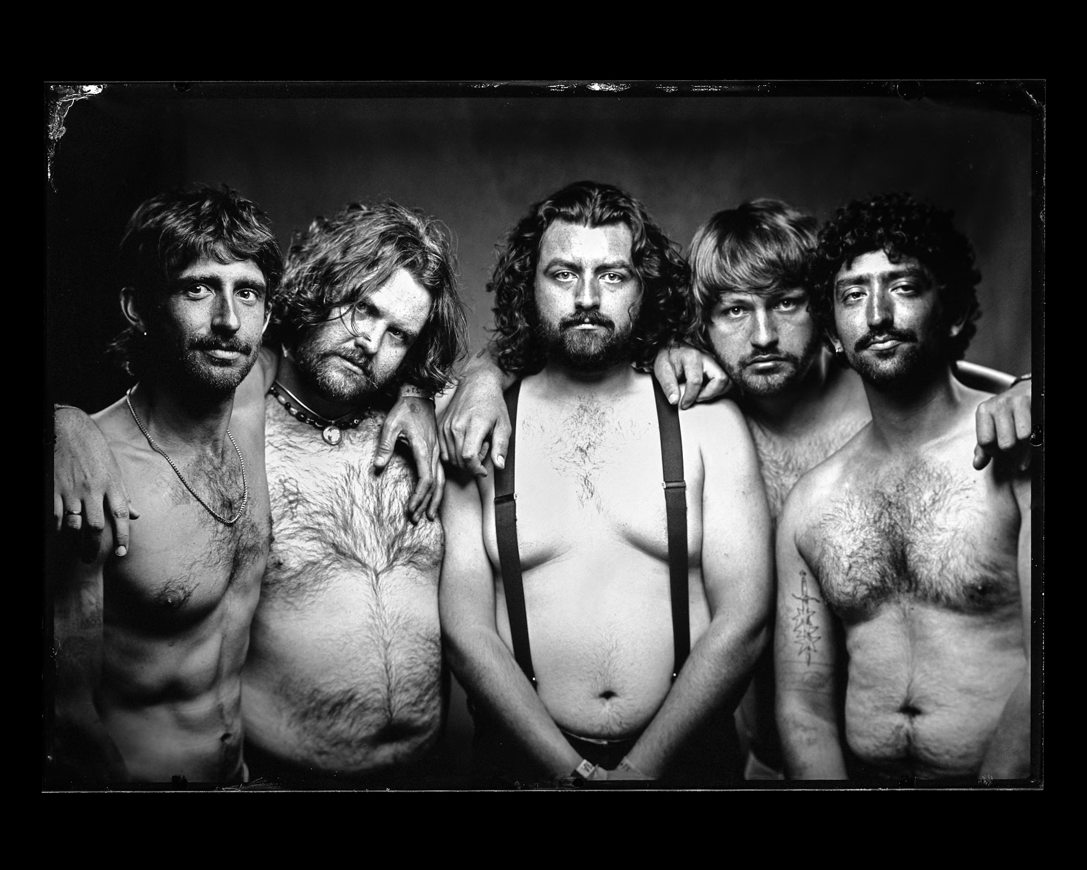 Five shirtless men with long hair and beards pose together, some with arms around each other, against a dark background.