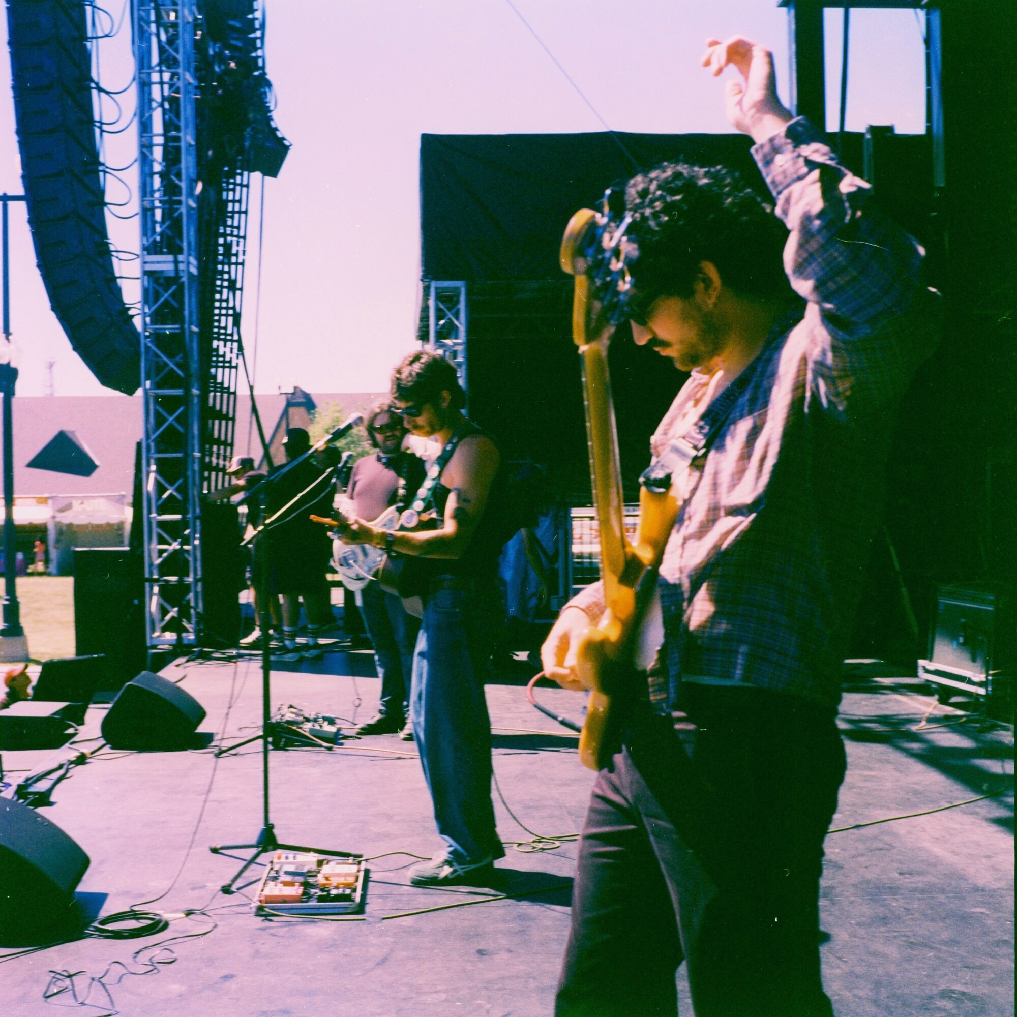 Musicians perform on an outdoor stage with instruments and microphones, under a clear sky, with stage equipment visible.