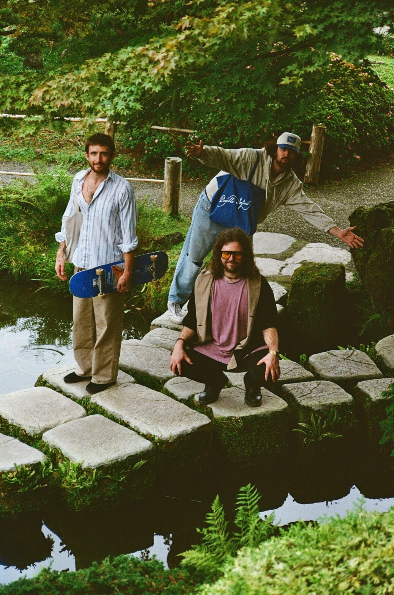 Three people on a stone bridge over a stream in a park, surrounded by green trees and plants.