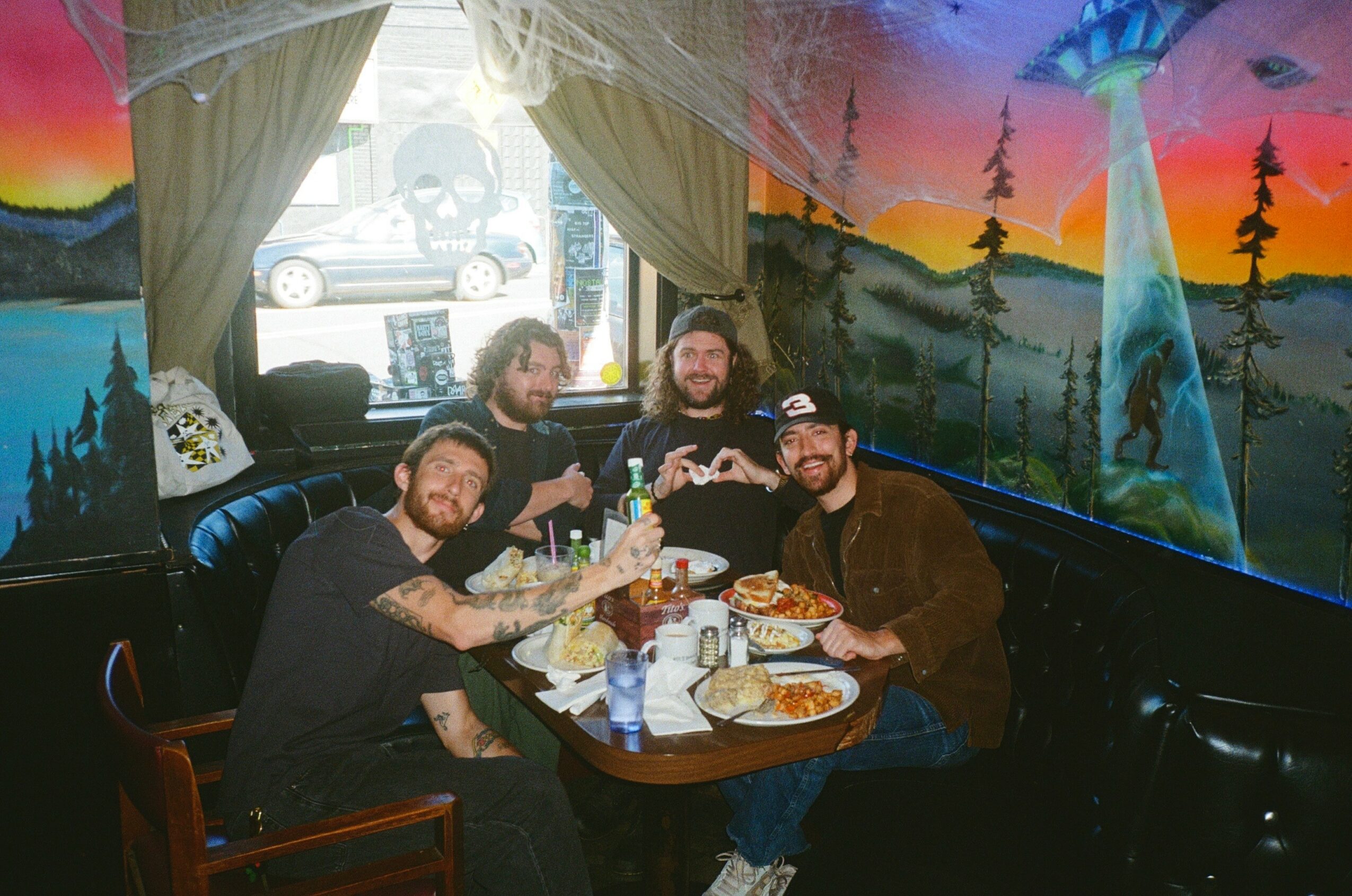 Four people sit around a table in a colorful, painted room with mountain and forest murals, enjoying food and drinks.