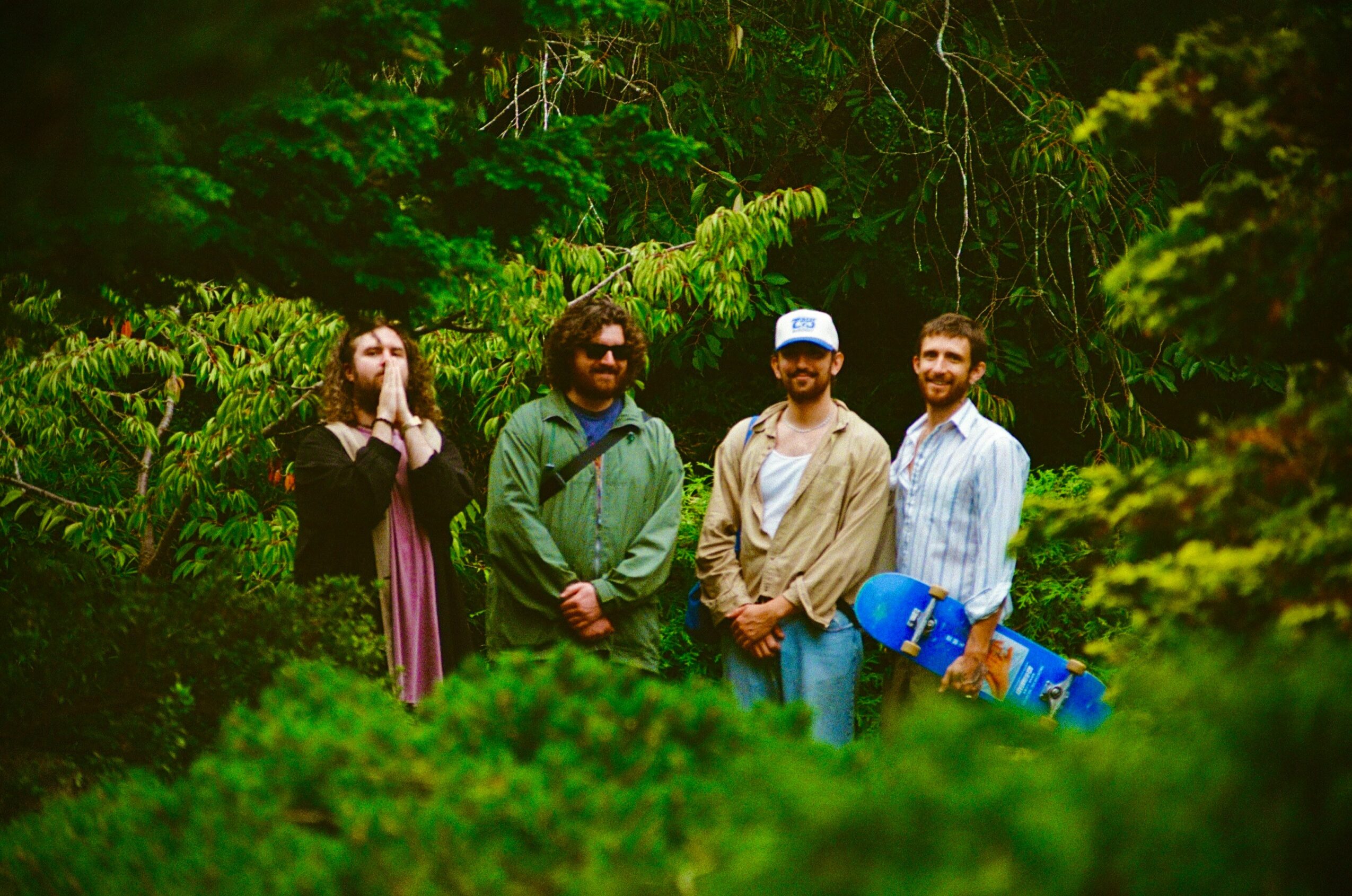 Four people standing outdoors among trees and greenery, with one holding a skateboard, smiling and posing for the photo.