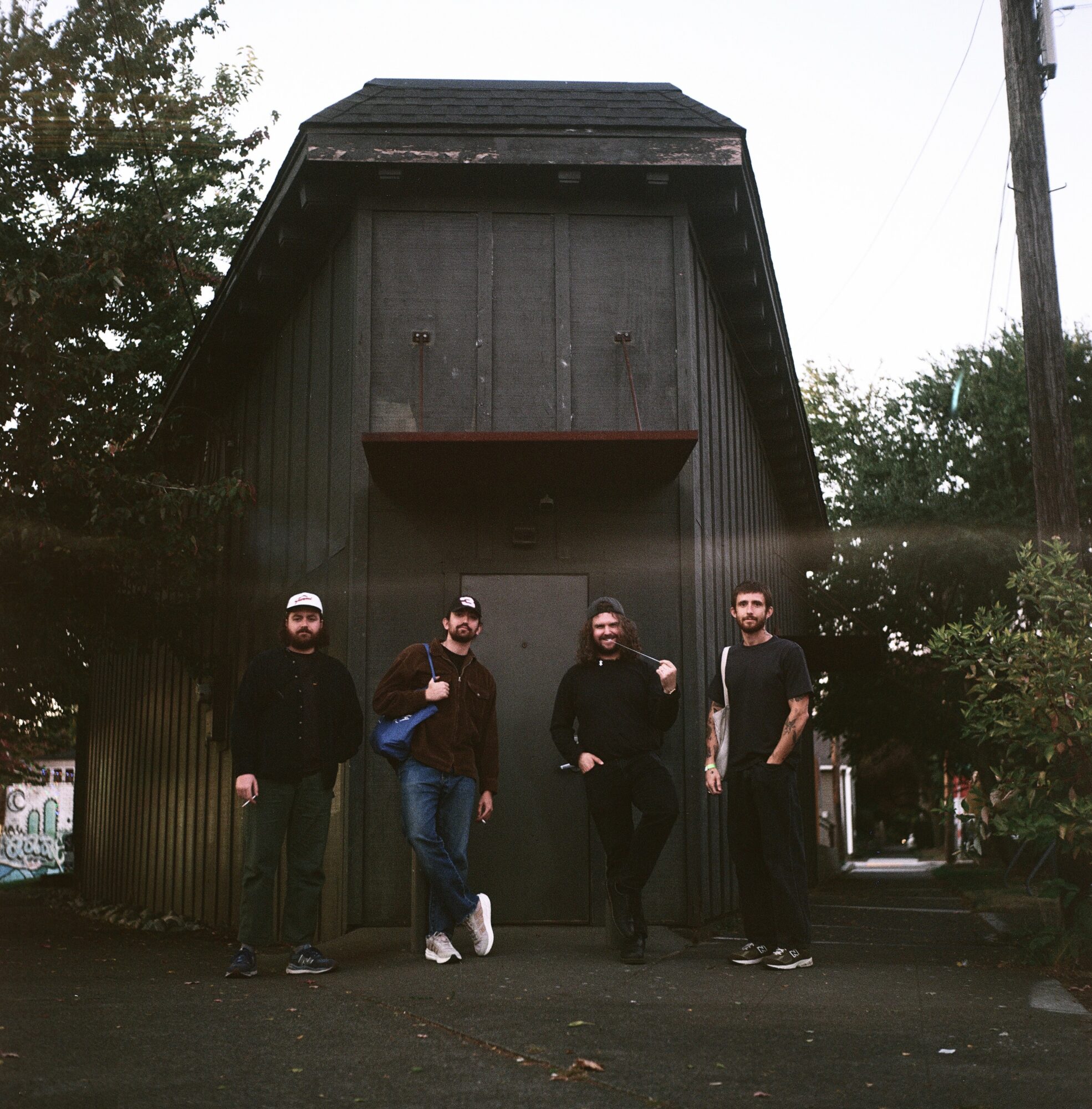 Four people standing in front of a large dark wooden building with trees and power lines in the background.