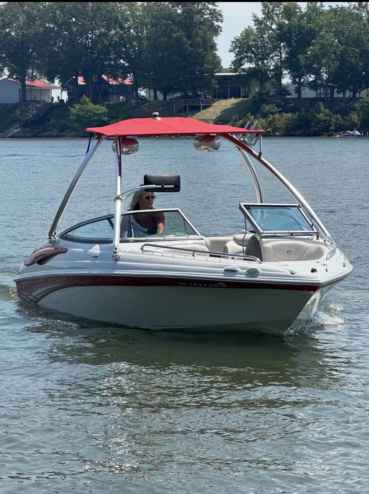 Motorboat with a red canopy on water, trees and buildings in background, person on boat steering, sunny day