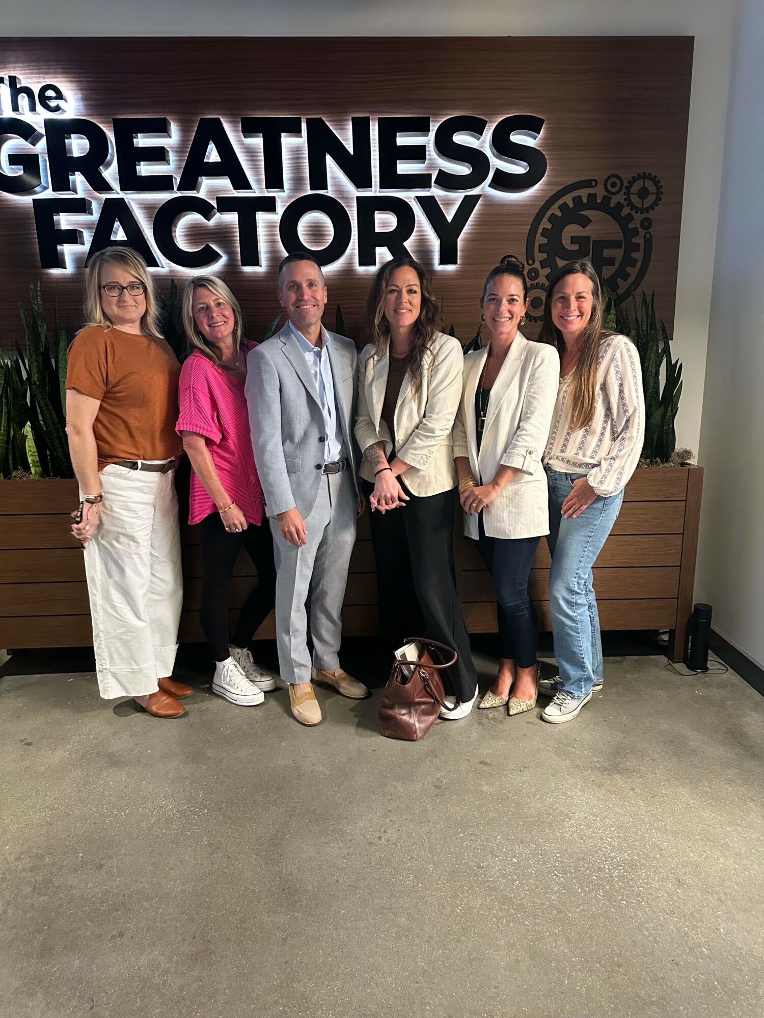 Group of seven people standing indoors, smiling, with a large sign reading 'The Greatness Factory' behind them.