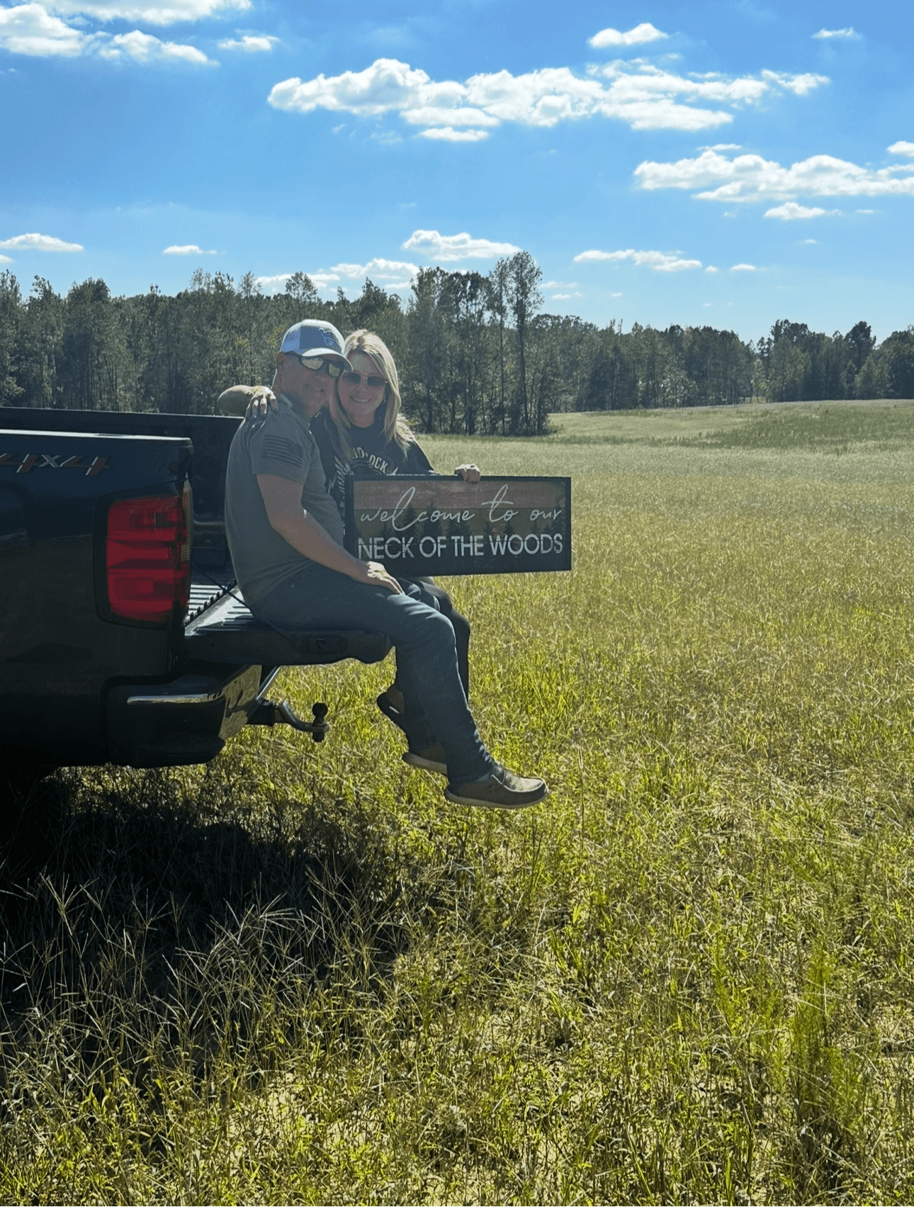 Two people sitting on the back of a truck in a grassy field, holding a sign that reads 'NECK OF THE WOODS,' with trees and a blue sky in the background.