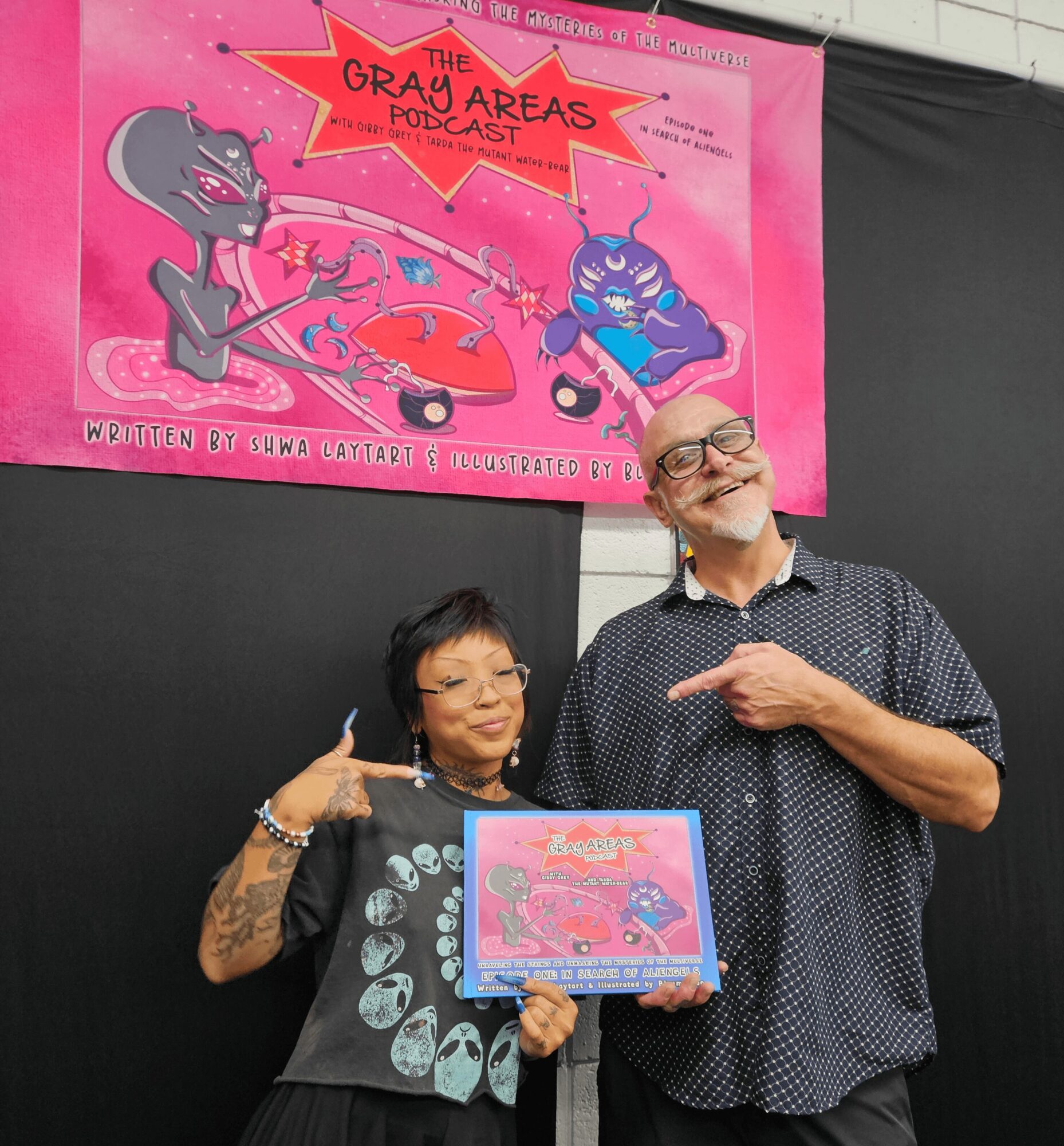 Two people holding a pink cartoon-themed poster and a framed picture, smiling, in front of a black background.