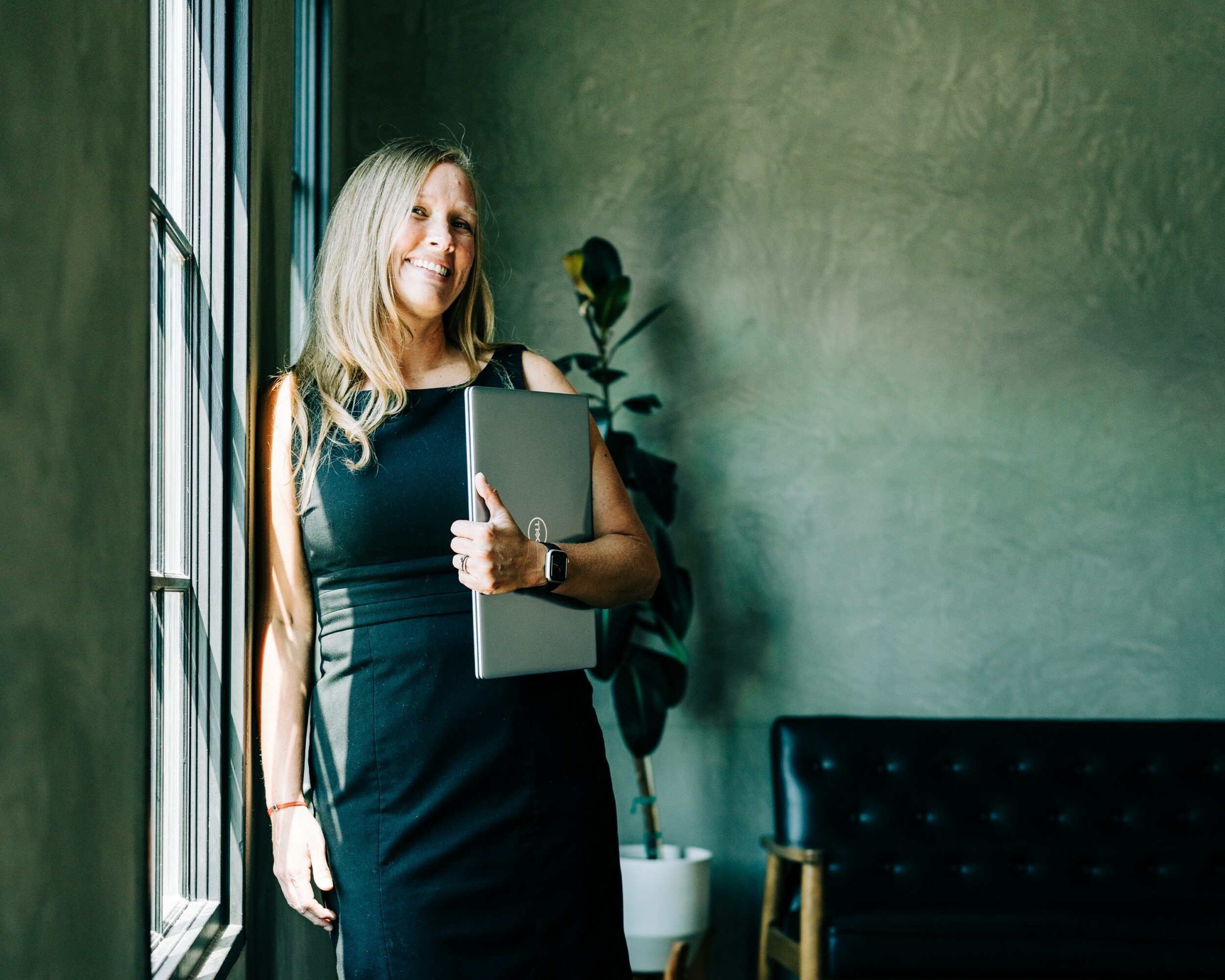Woman with long blonde hair holding a laptop, standing near a window with a green wall background.