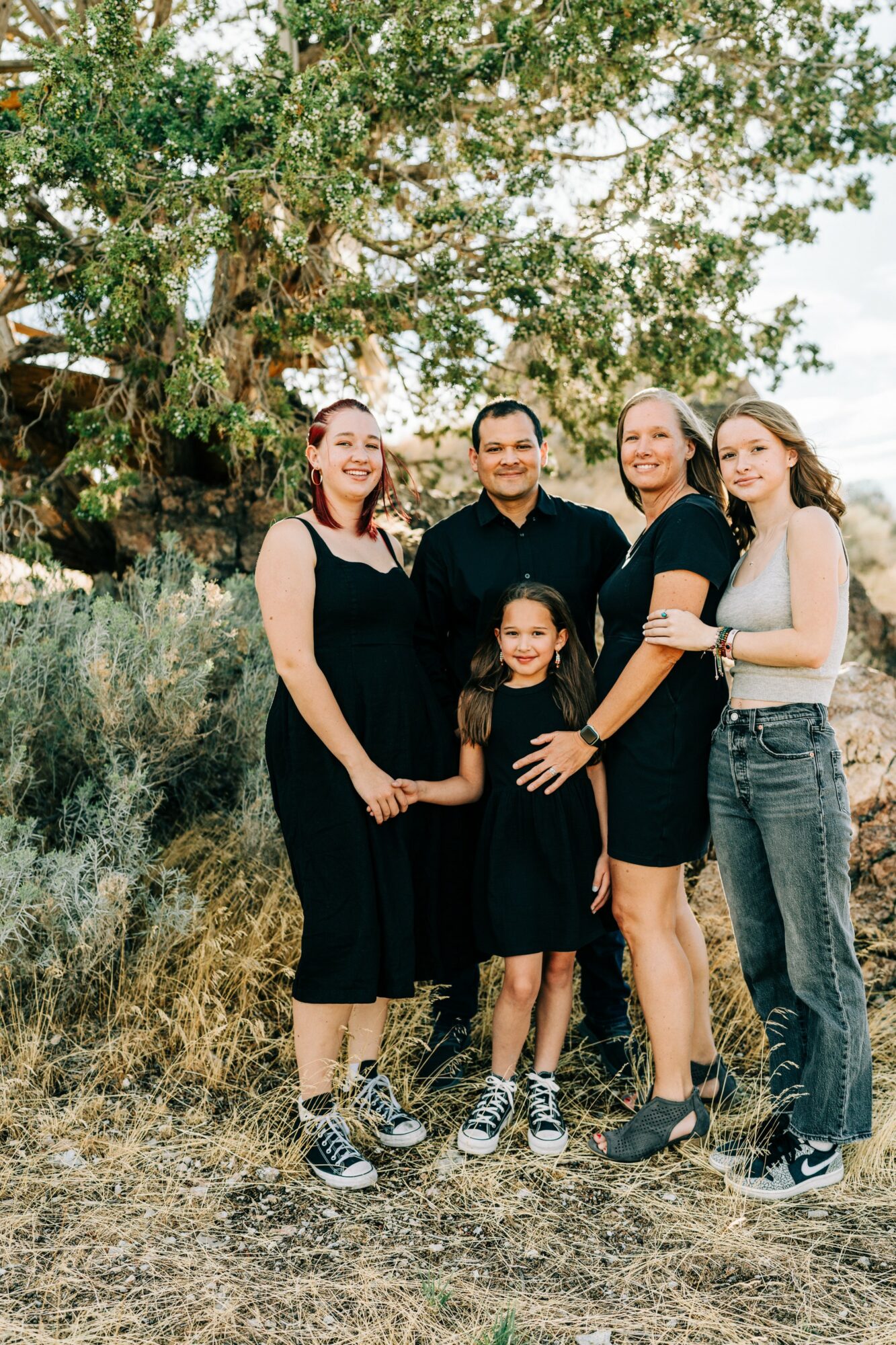 Group of five people, including children, standing outdoors in front of a large tree and greenery, smiling at the camera.