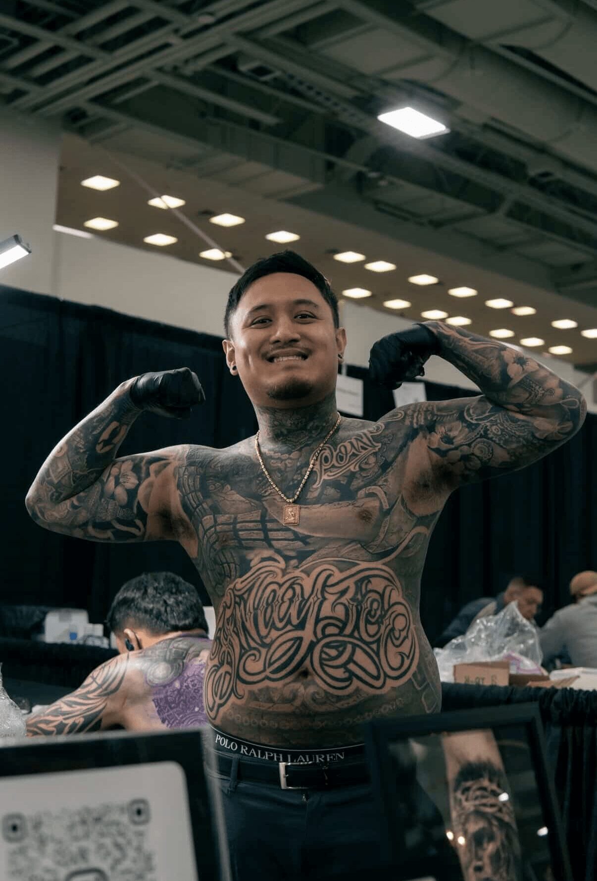 Young man with tattoos flexing his muscles in an indoor event space, smiling, with people and black curtains in background.
