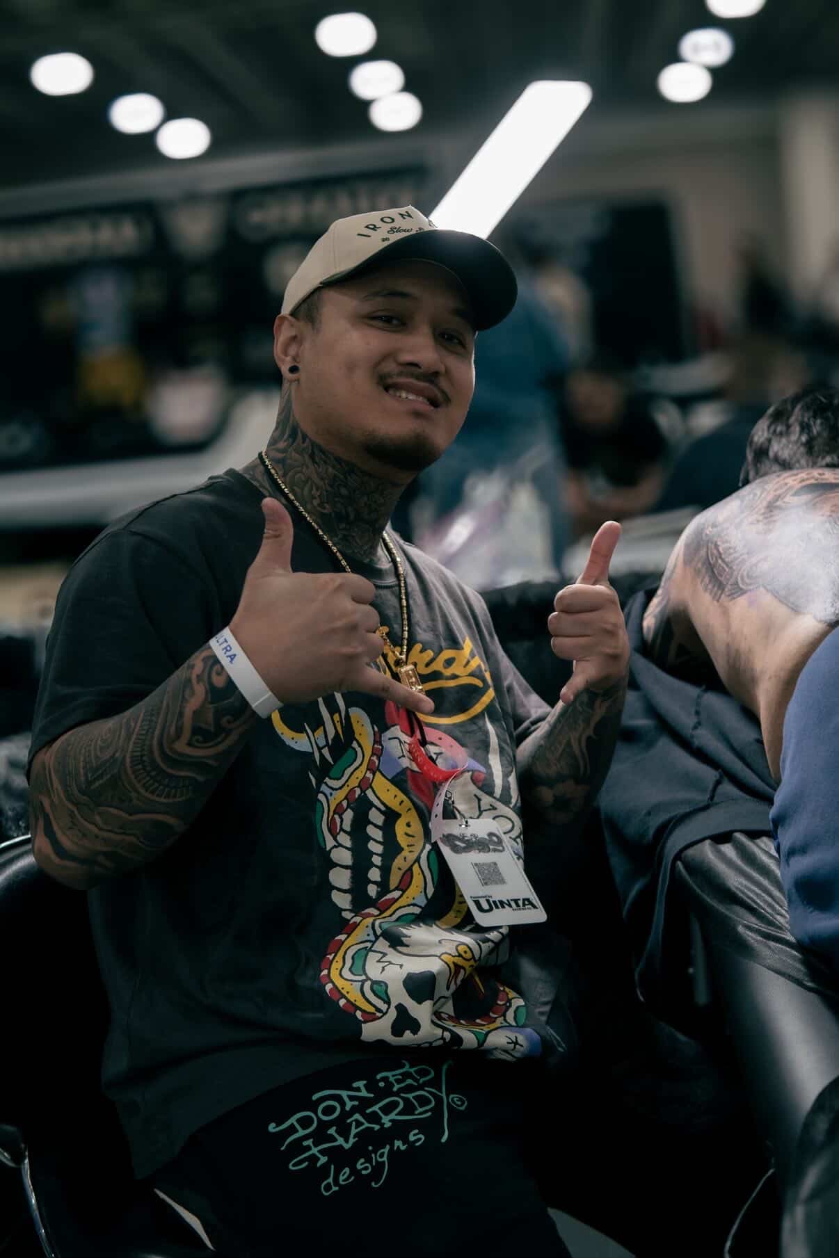 Young man with tattoos wearing a cap and black shirt, smiling and giving thumbs-up at an indoor event.