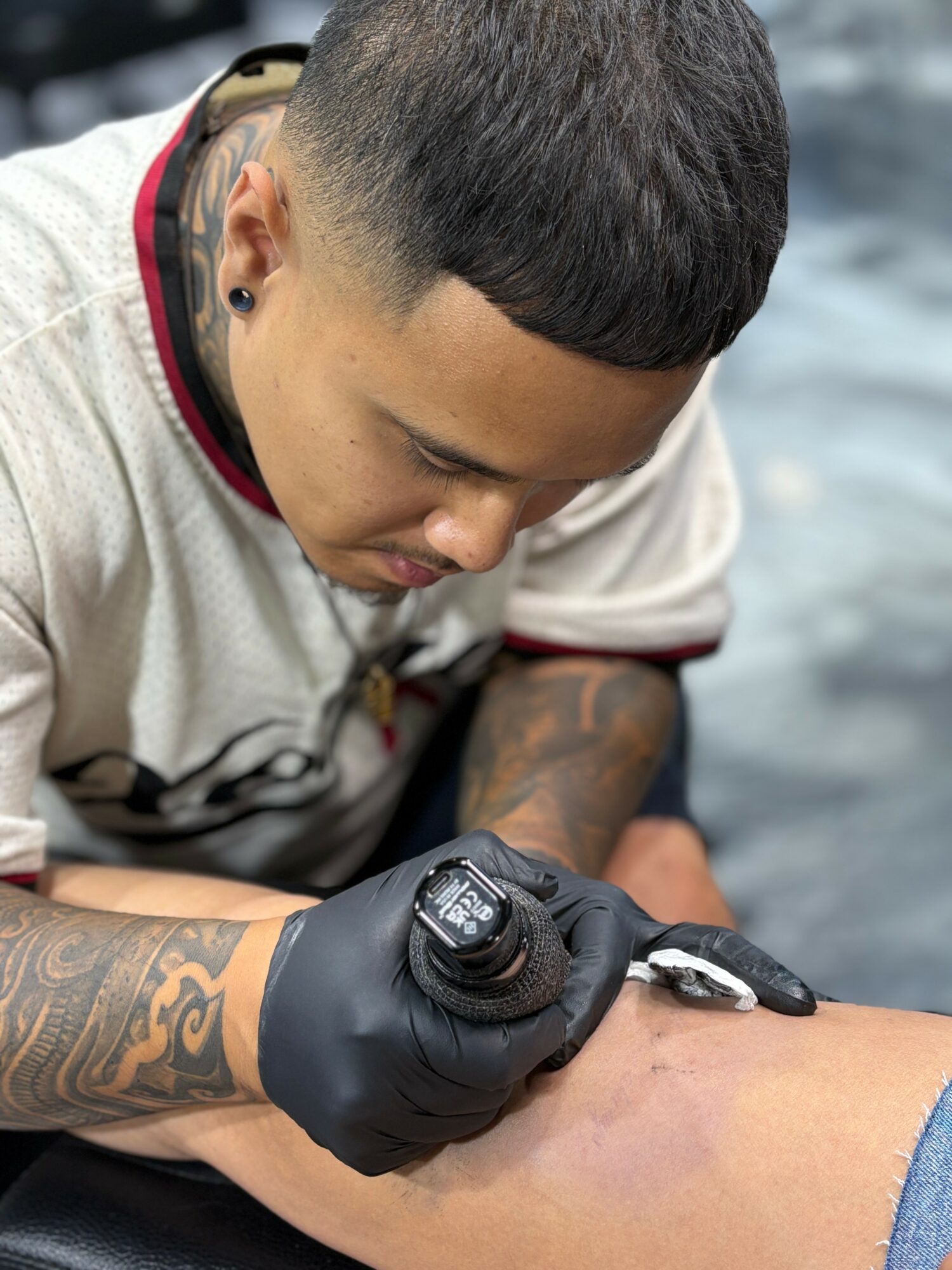 Person with short hair and tattoos on arm, wearing black glove, using tattoo machine on skin, focused downward.