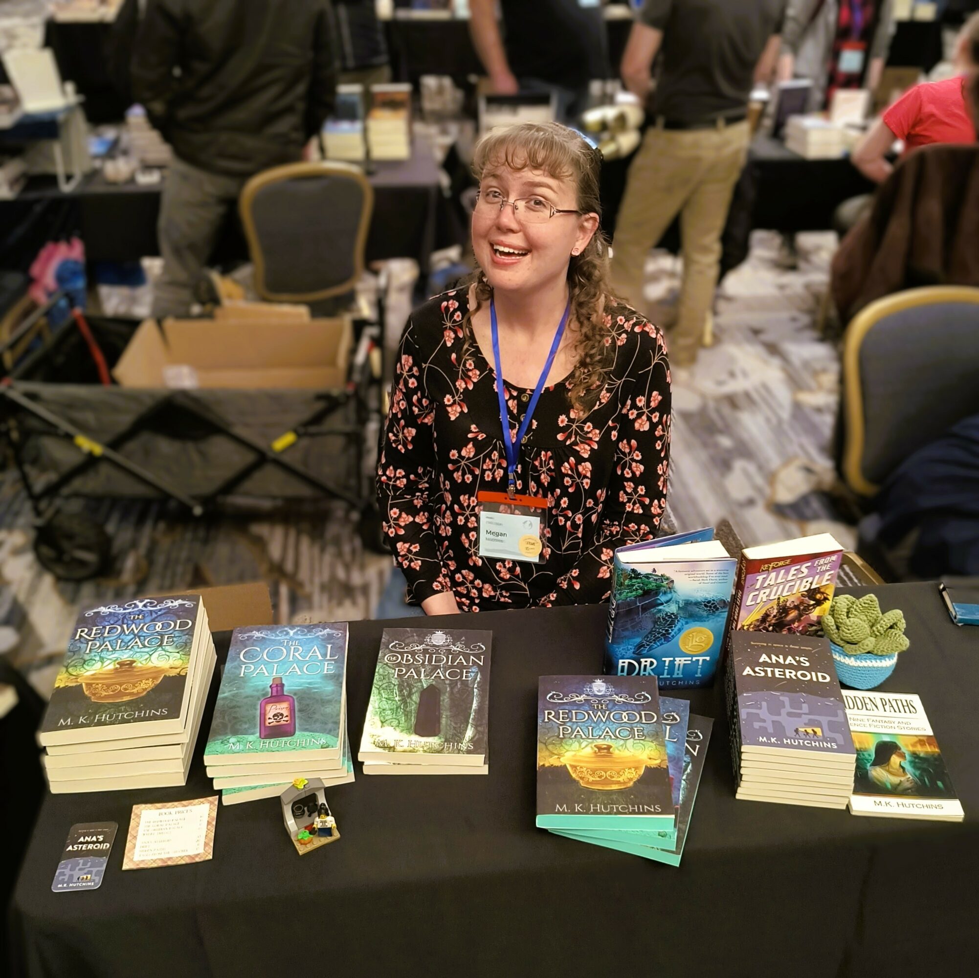 Young woman with glasses and a floral dress sits behind a table with books at a convention, smiling. People are in the background.