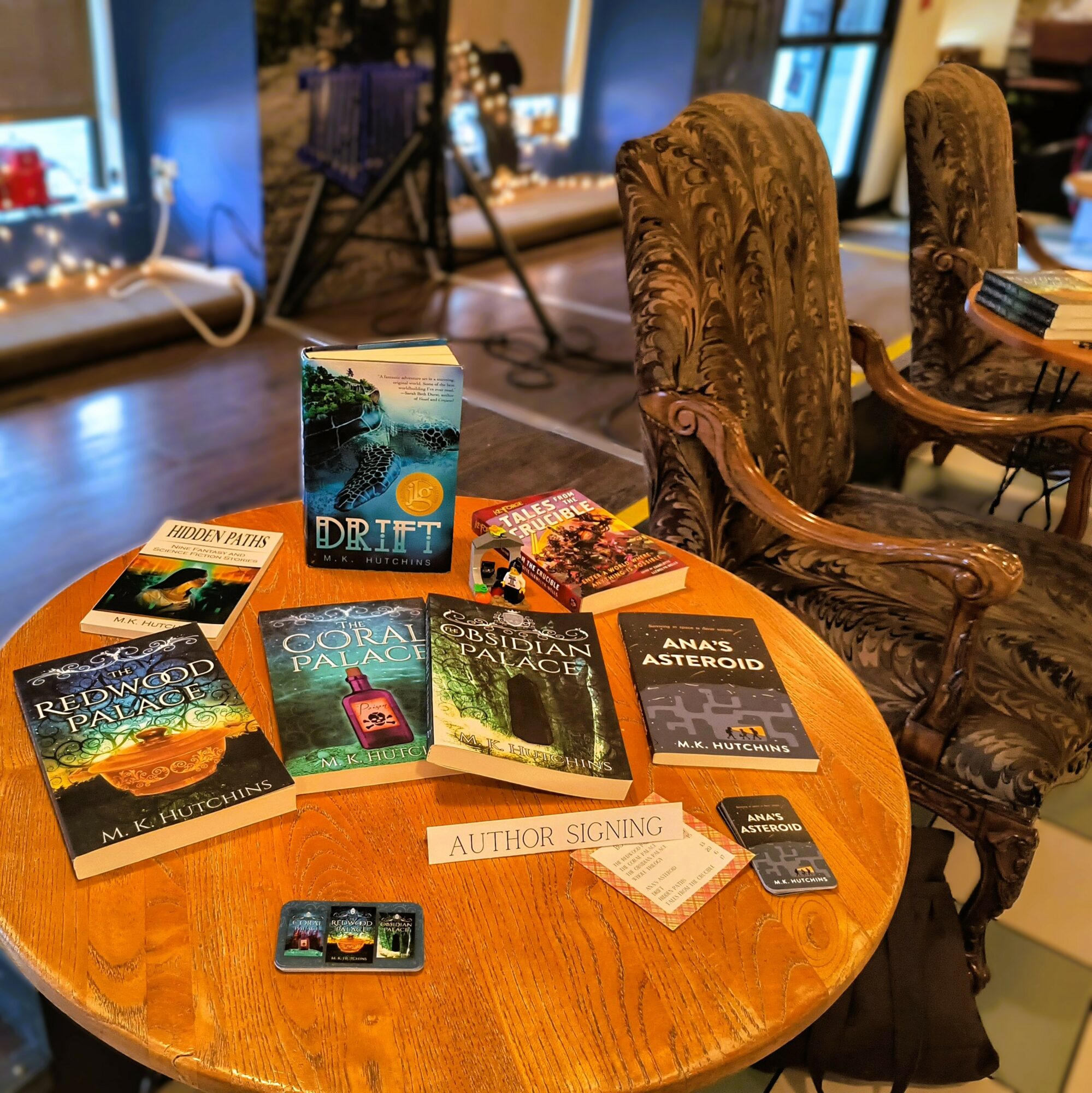 Round wooden table with books, a sign for author signing, and a small device, in a cozy room with ornate chairs.