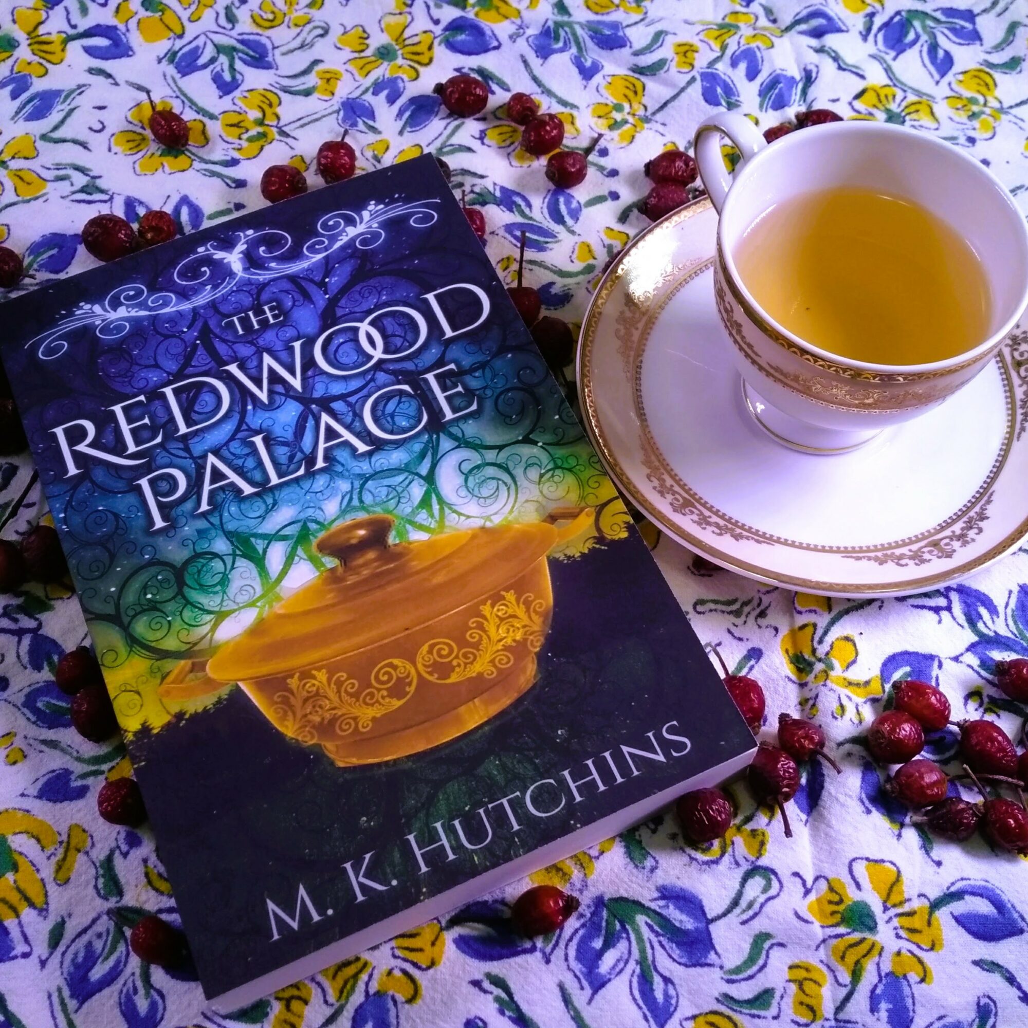 Book titled 'The Redwood Palace' by M. K. Hutchins, a teacup with tea on a floral tablecloth, surrounded by cherries.