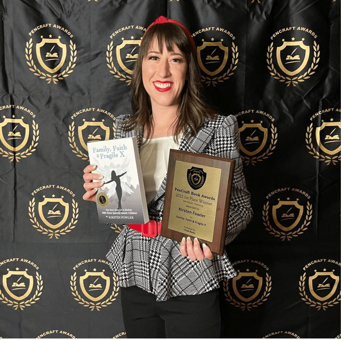 Woman holding a book and a plaque, standing in front of a black backdrop with gold logos, smiling, wearing a checkered blazer.