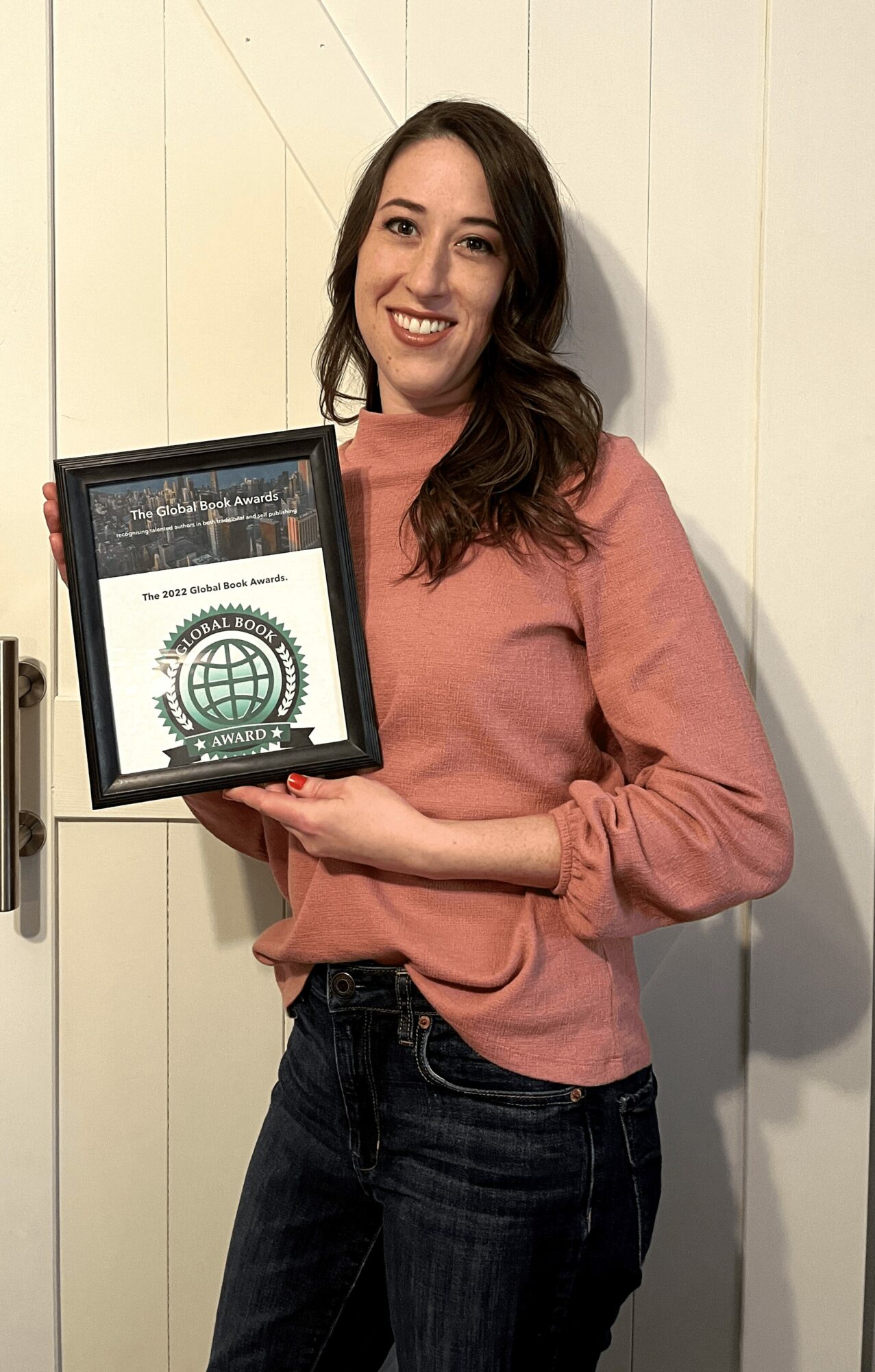 Woman with long dark hair smiling, holding a framed certificate or award, standing against a white wall.