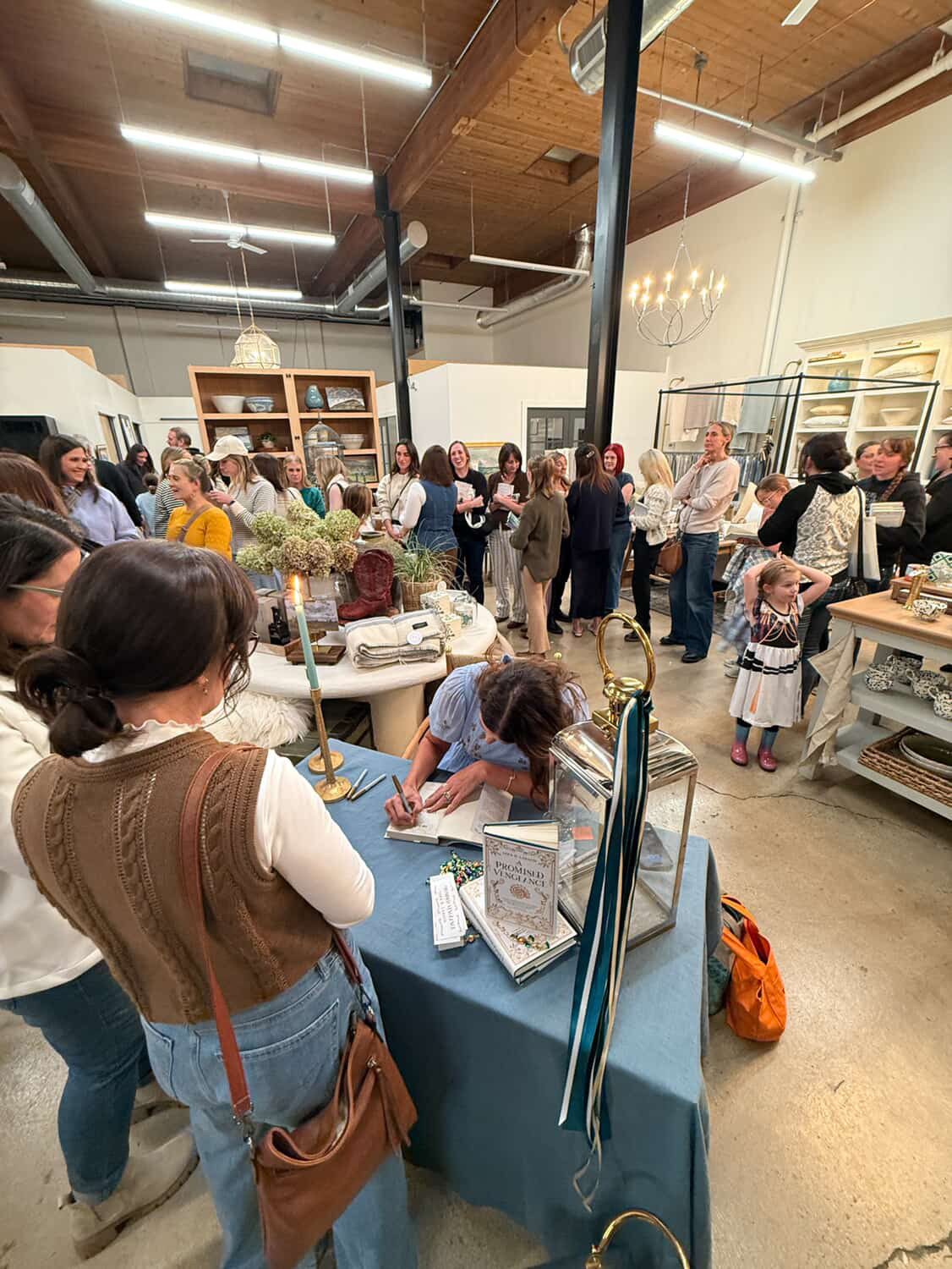 People gather around tables and displays in a spacious indoor market or craft fair with high ceilings and wooden beams.
