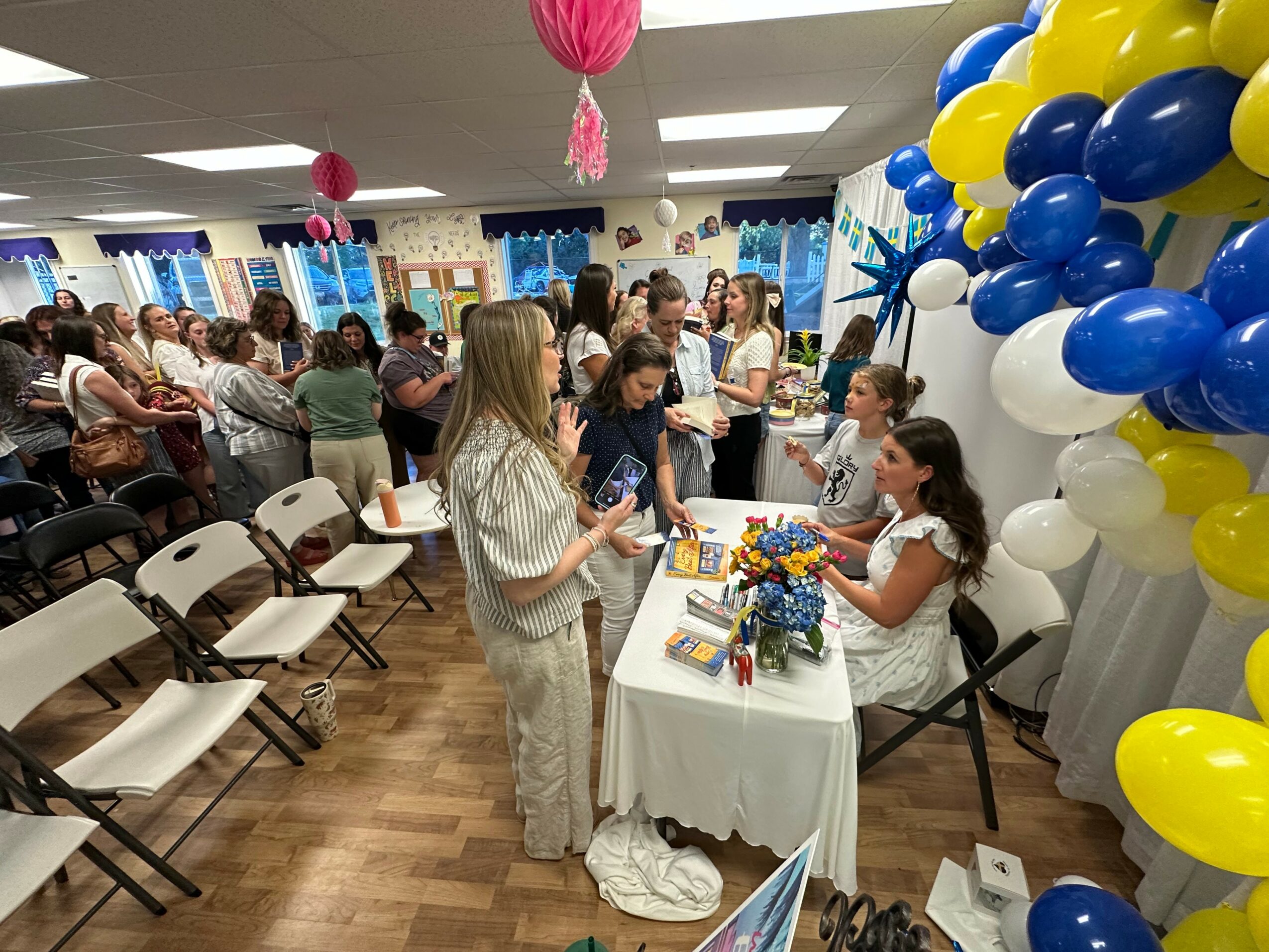 Children and adults gather around a table with balloons and decorations at a party, with chairs and people in the background.