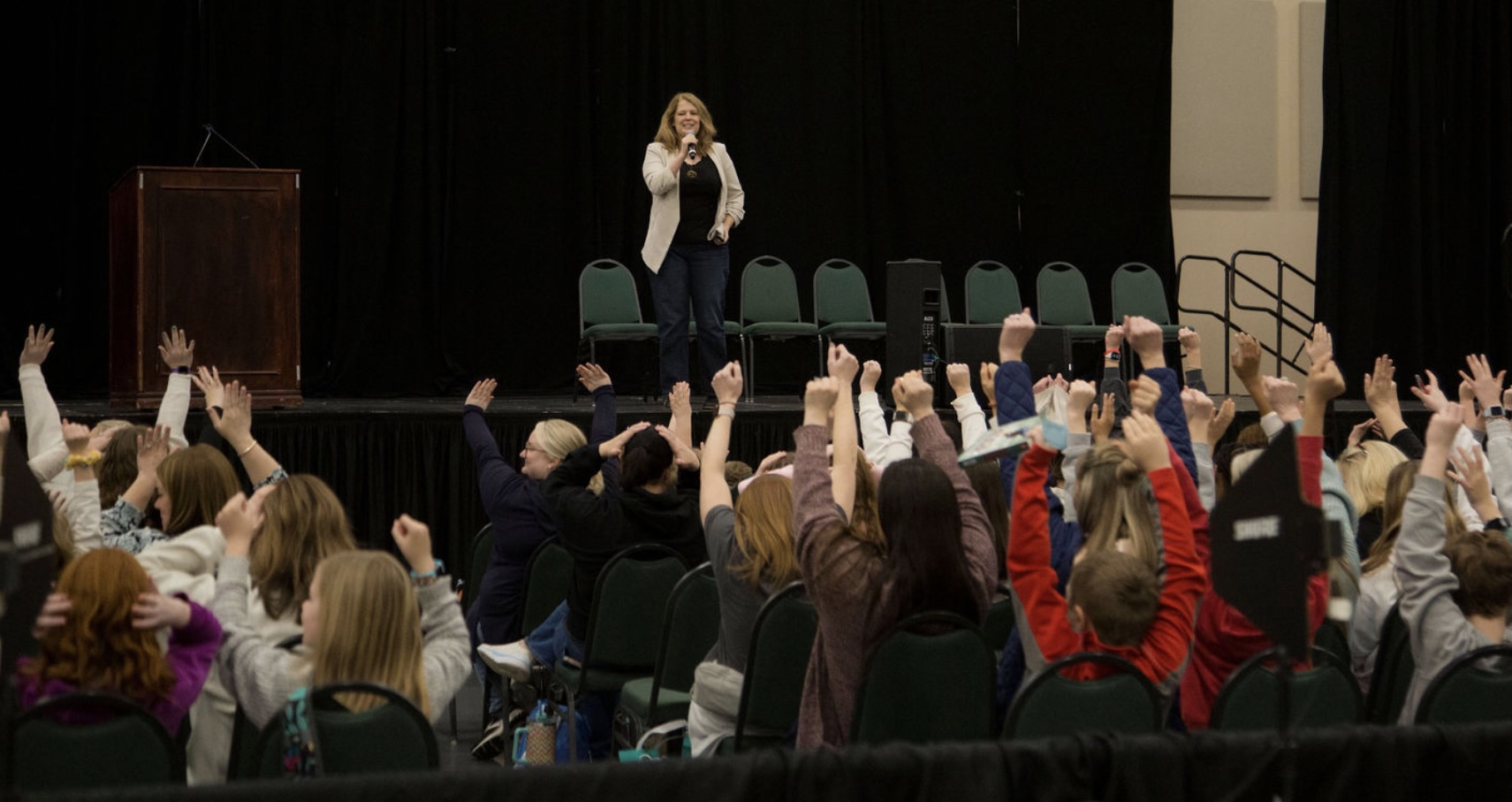 Person standing on stage speaking to audience with raised hands, audience members raising hands, black curtains backdrop.