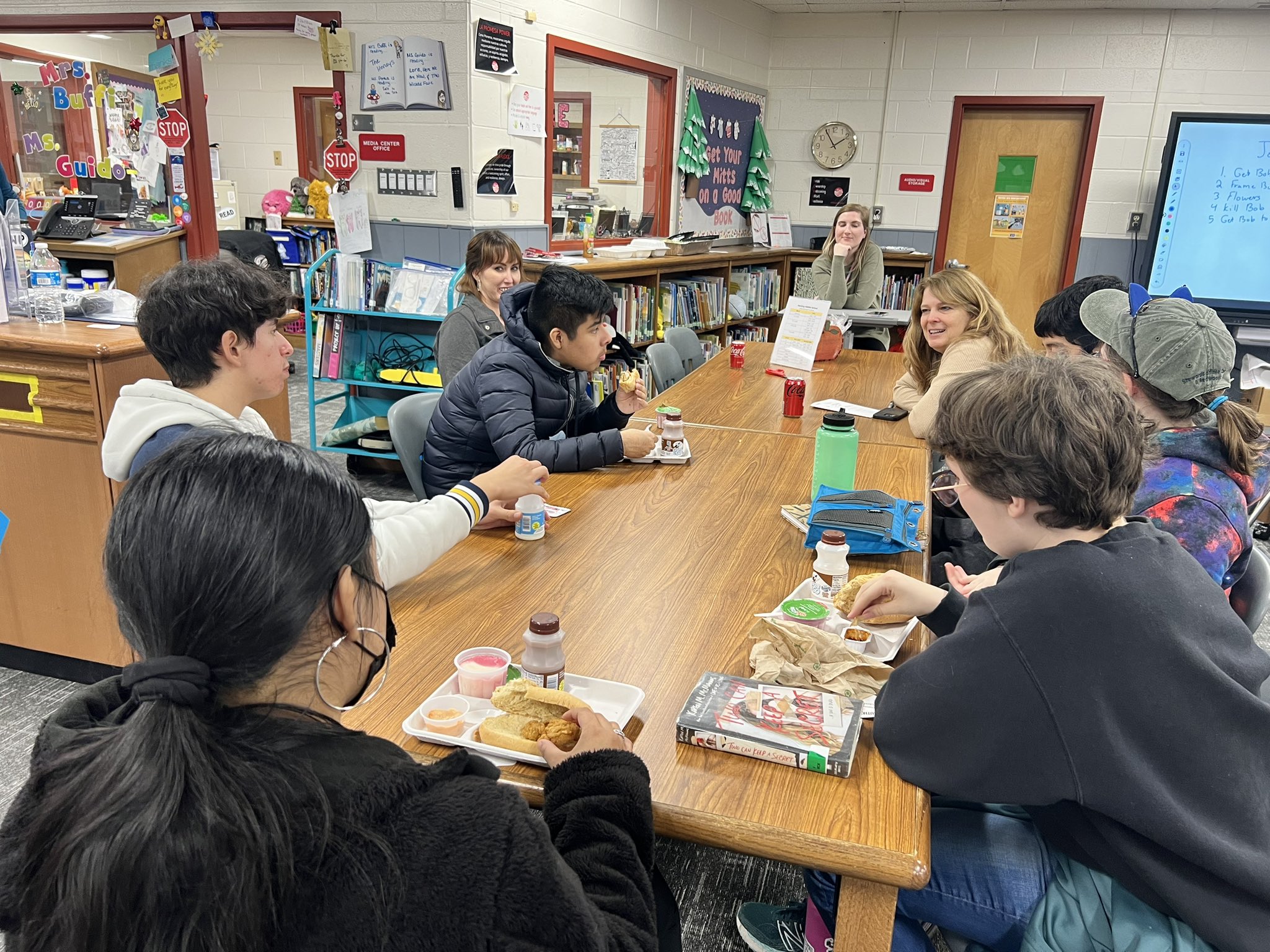 Group of students sitting around a classroom table, eating snacks and talking, with bookshelves and classroom decorations in the background.