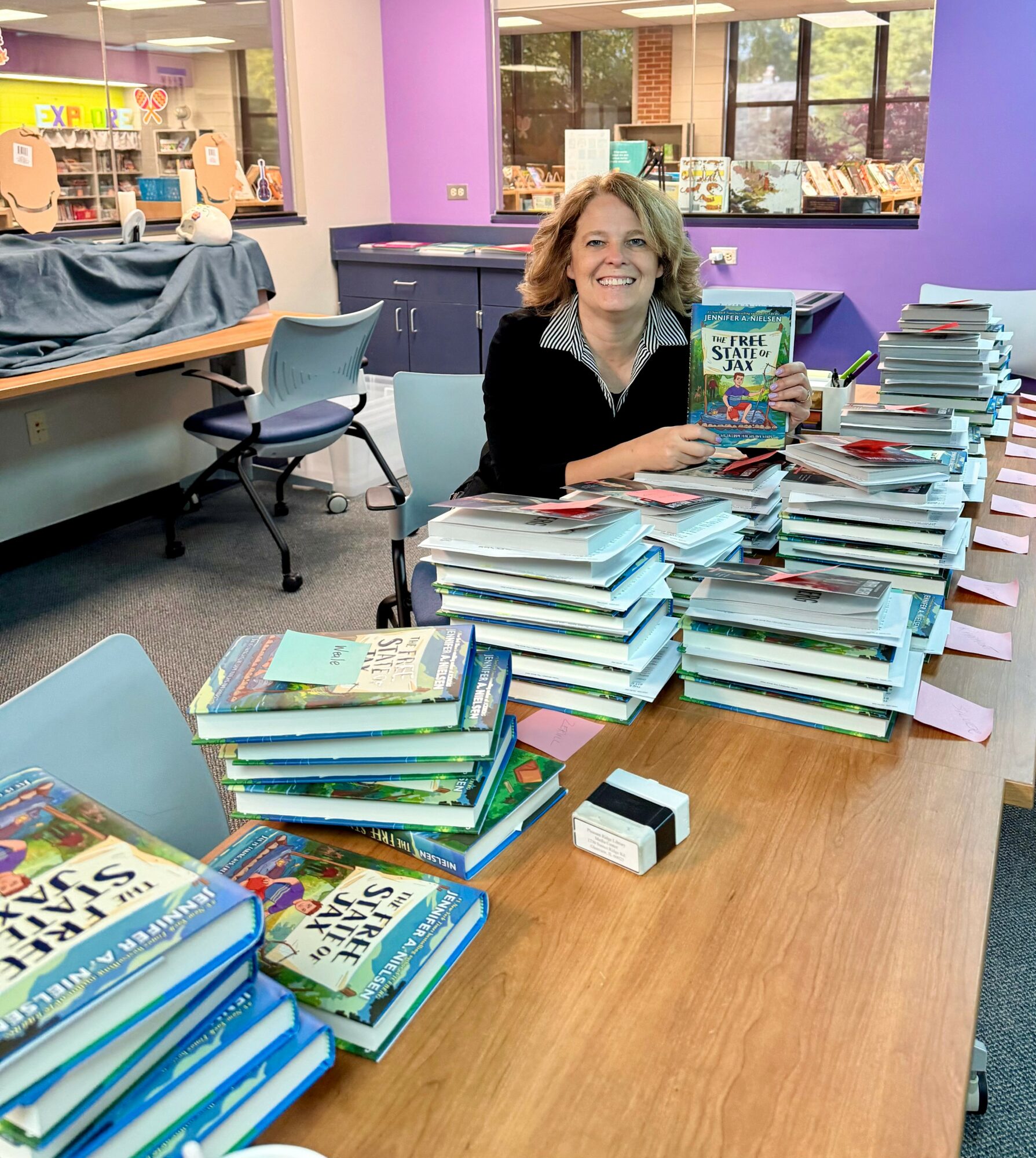 Woman smiling at a table with stacks of books in a colorful library or classroom setting.