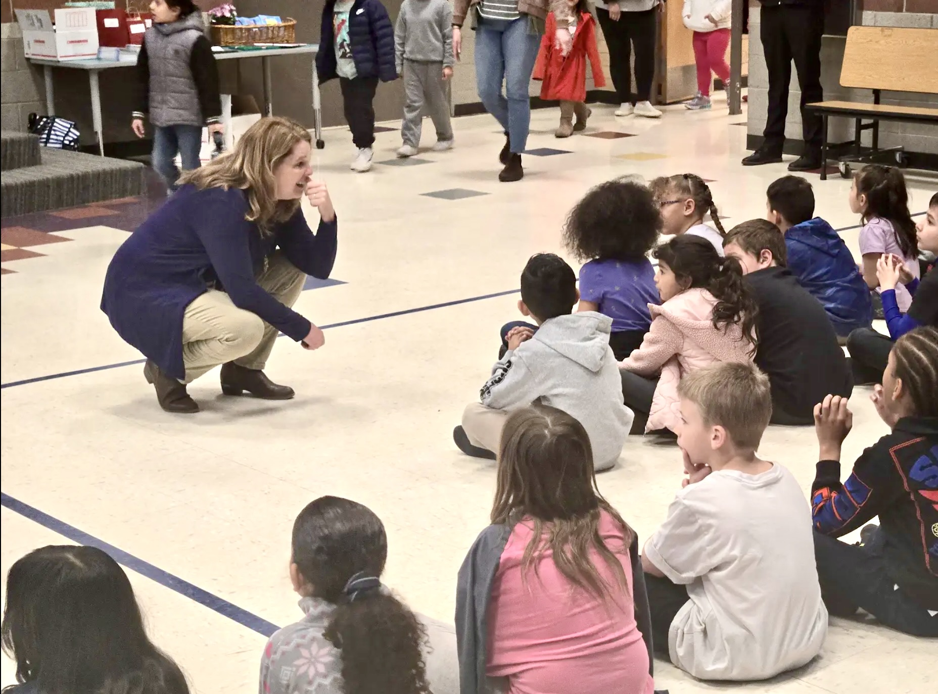 A woman crouches and speaks to a group of children sitting on the floor in a public indoor space.