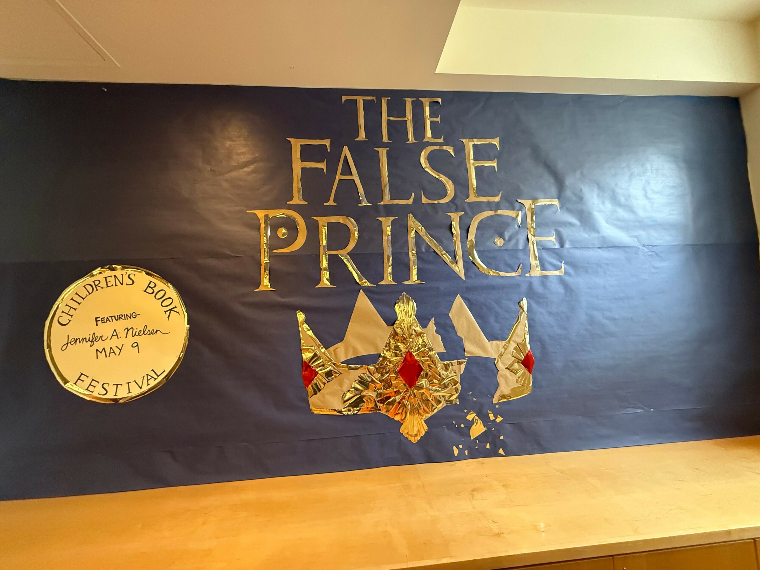 Wall with a dark background and gold text reading 'The False Prince,' and a decorative crown with red and gold details. A round yellow sign on the left says 'Children's Book Festival, May 4.'
