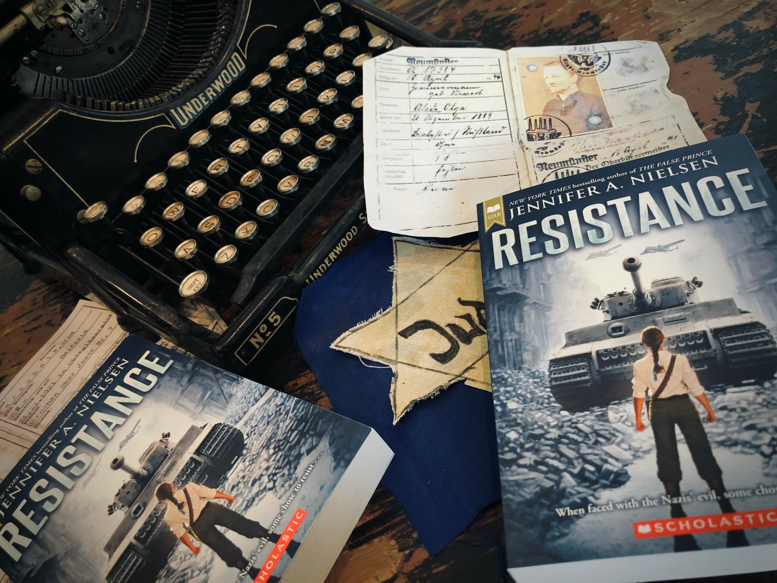 Books titled 'Resistance' and a notebook with handwritten notes on a wooden table, with a typewriter nearby.