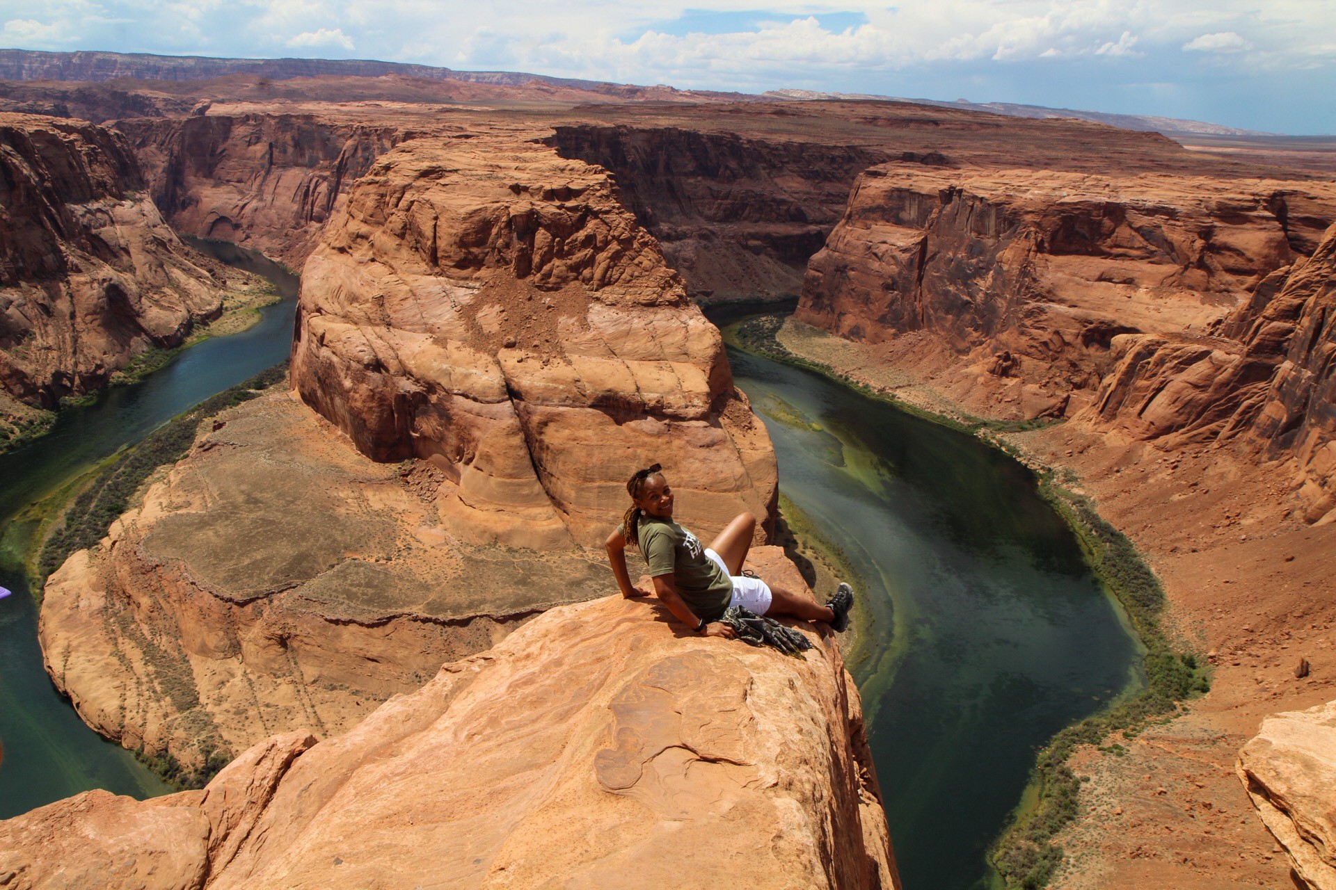Person sitting on edge of canyon overlooking a river, with cliffs and sky in background.
