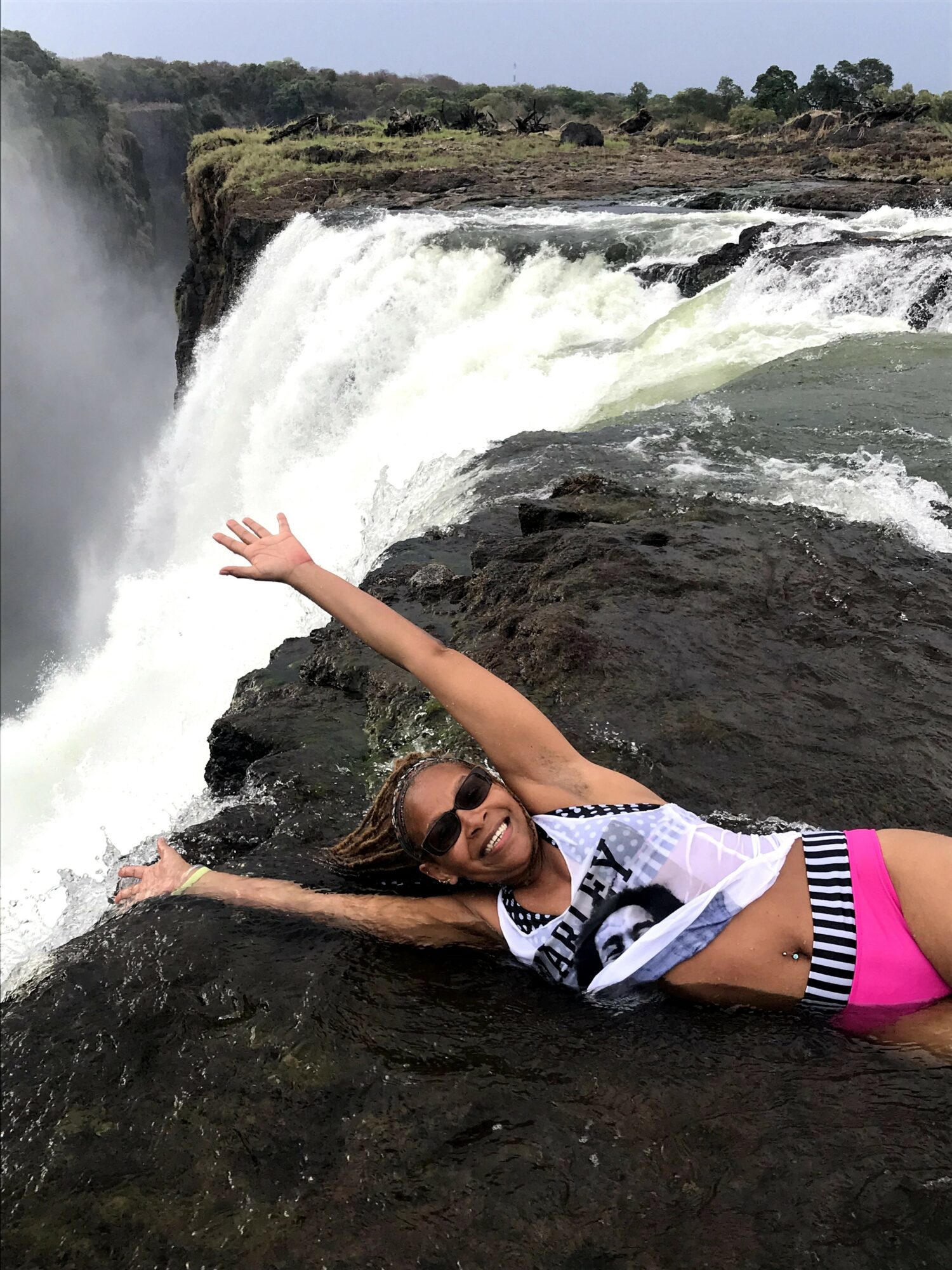Person lying on rocks near waterfall, arms outstretched, wearing sunglasses and a striped shirt, with water and cliffs in background.