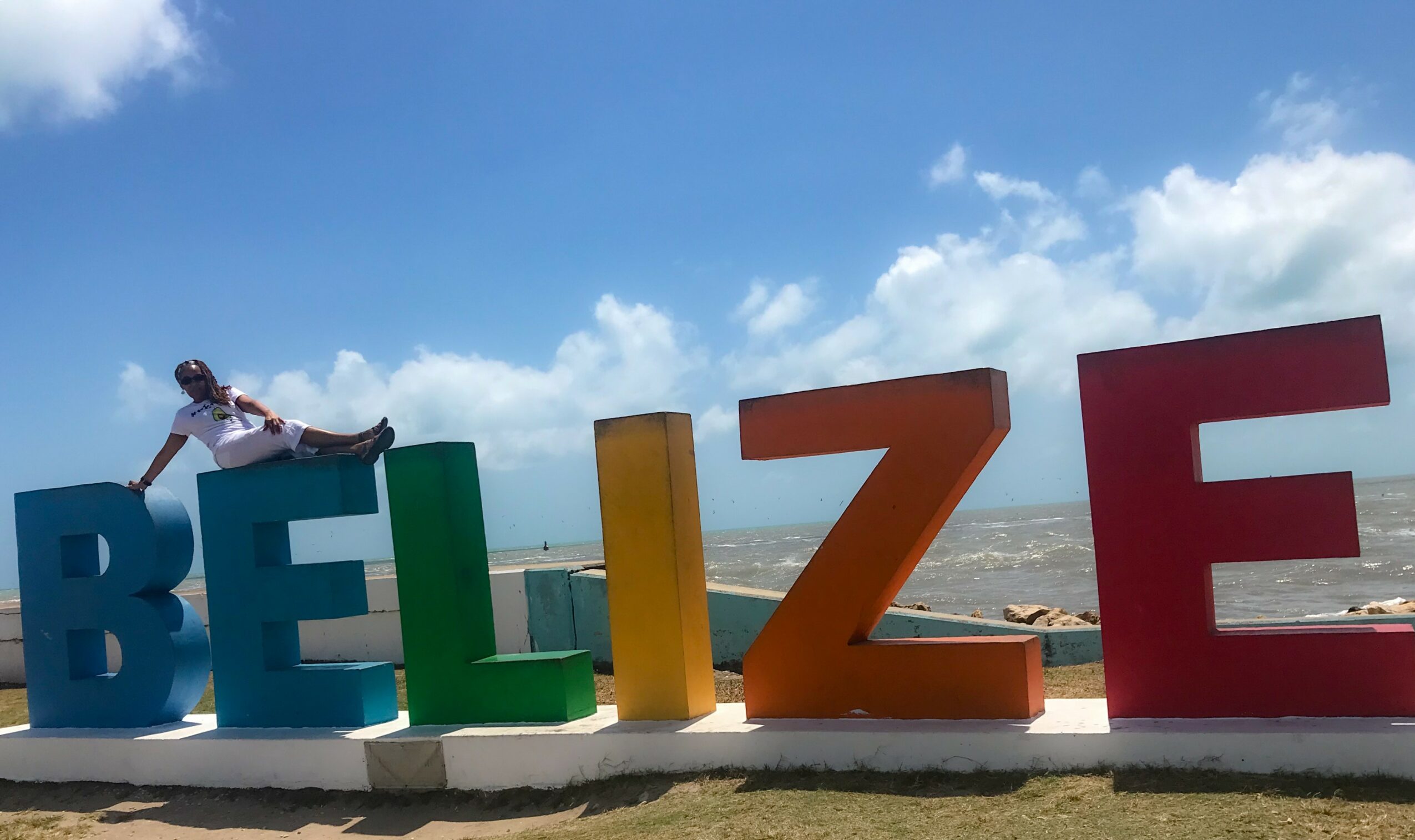 Person sitting on colorful letters spelling 'BELIZE' against a blue sky with clouds.
