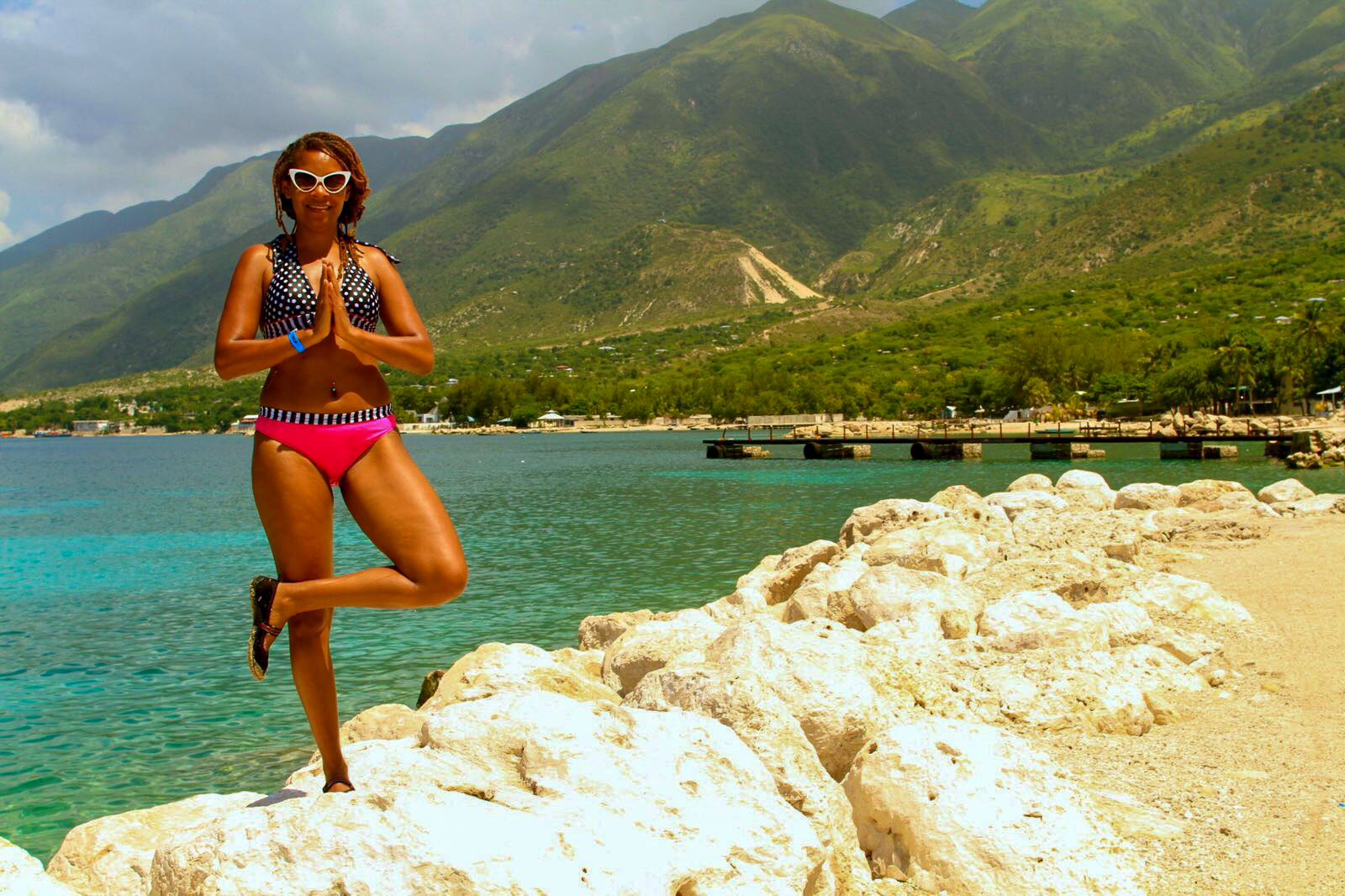 Woman in sunglasses and swimsuit practicing yoga on rocky beach with mountains and water in background.