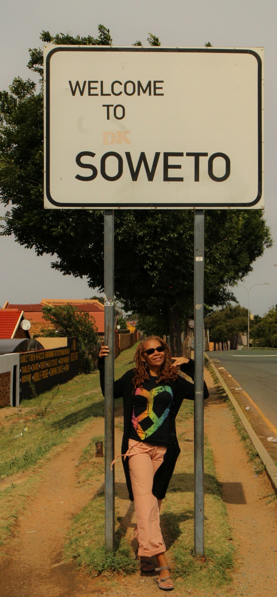 Person standing next to a welcome sign for Soweto, smiling, with trees and a road in the background.
