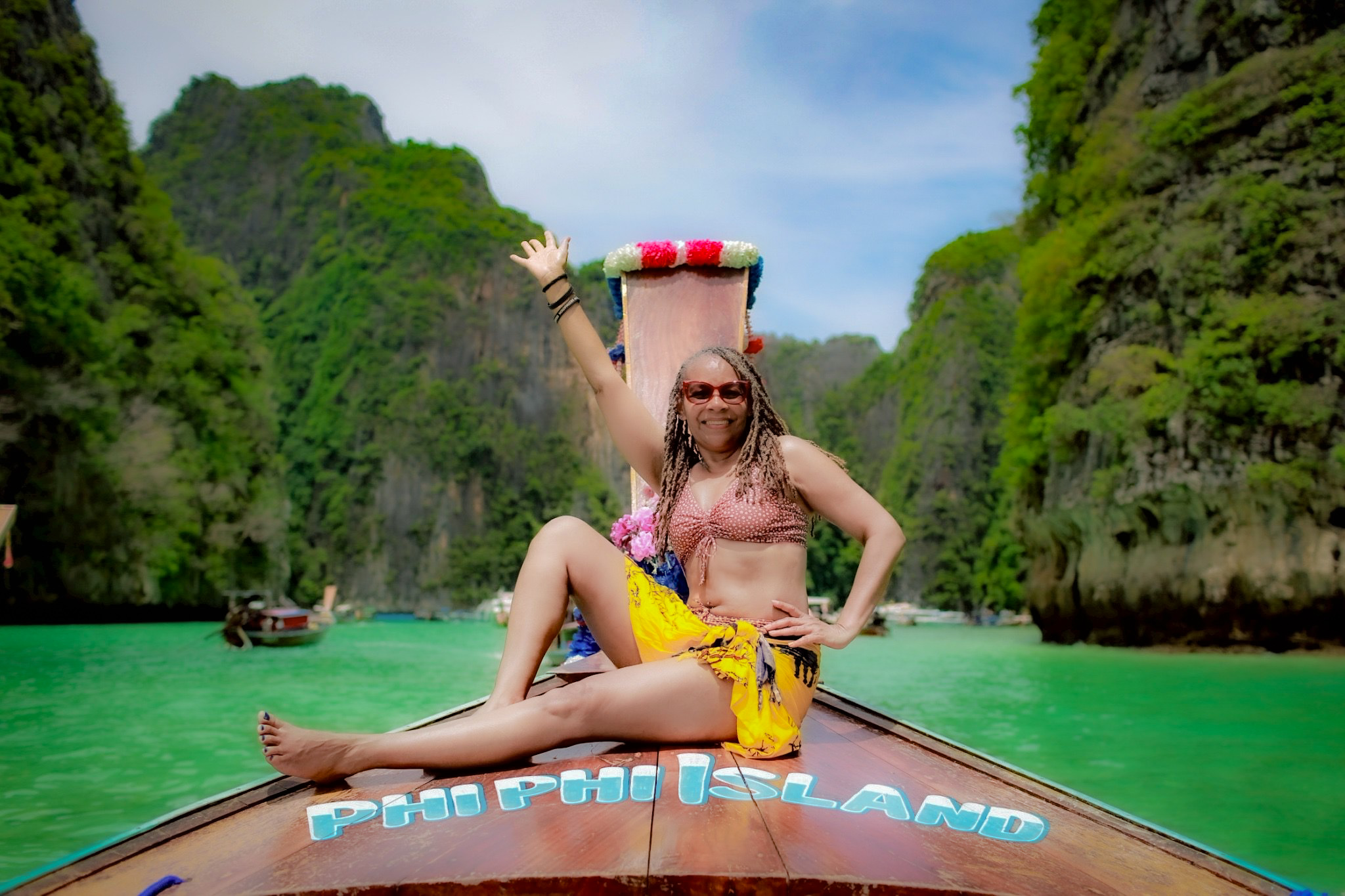 Woman sitting on boat with arms raised, surrounded by green cliffs and turquoise water, smiling, wearing sunglasses and colorful clothing.