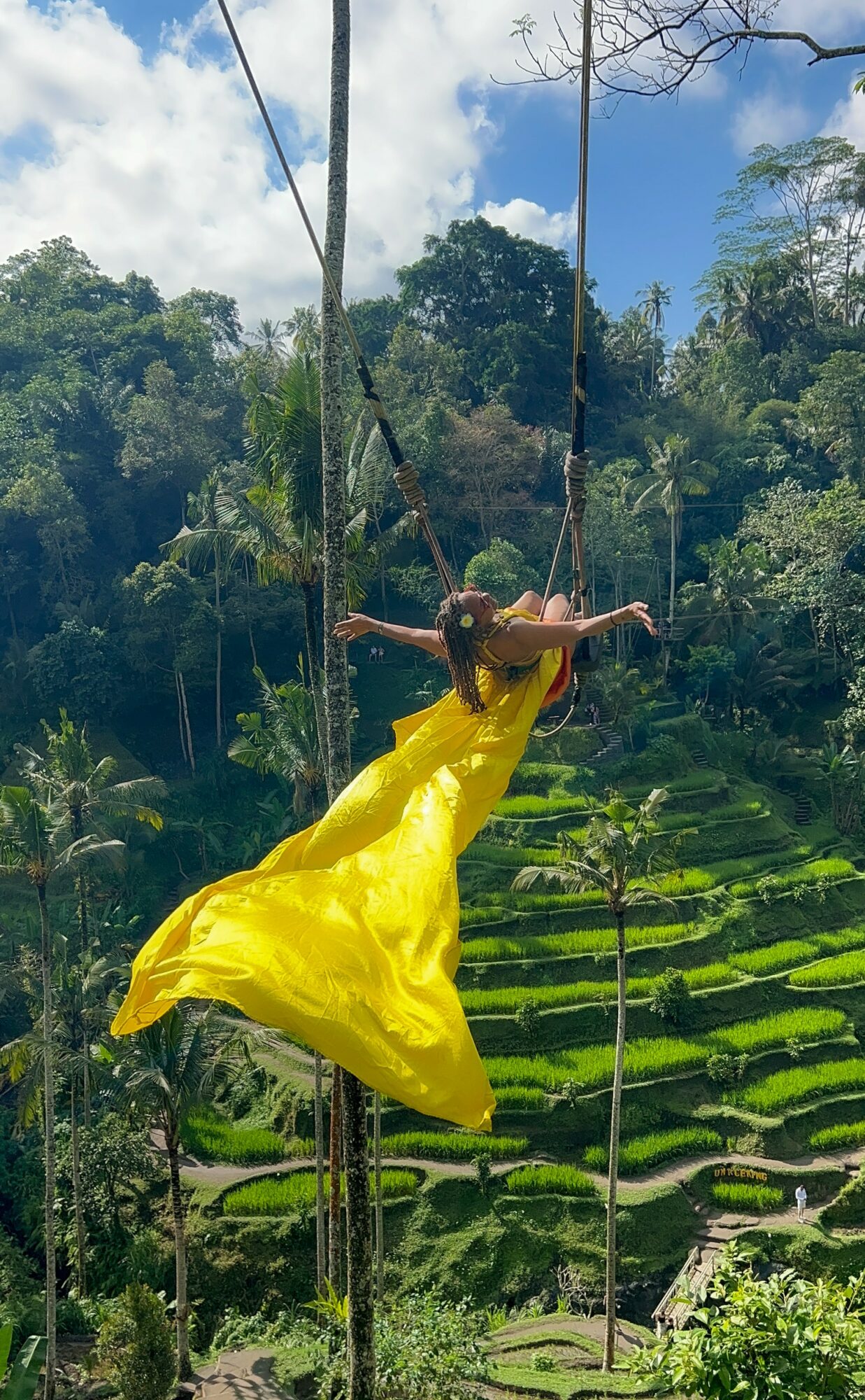 Person in a yellow dress swinging on a rope swing over green terraced fields and trees, blue sky with clouds in background.