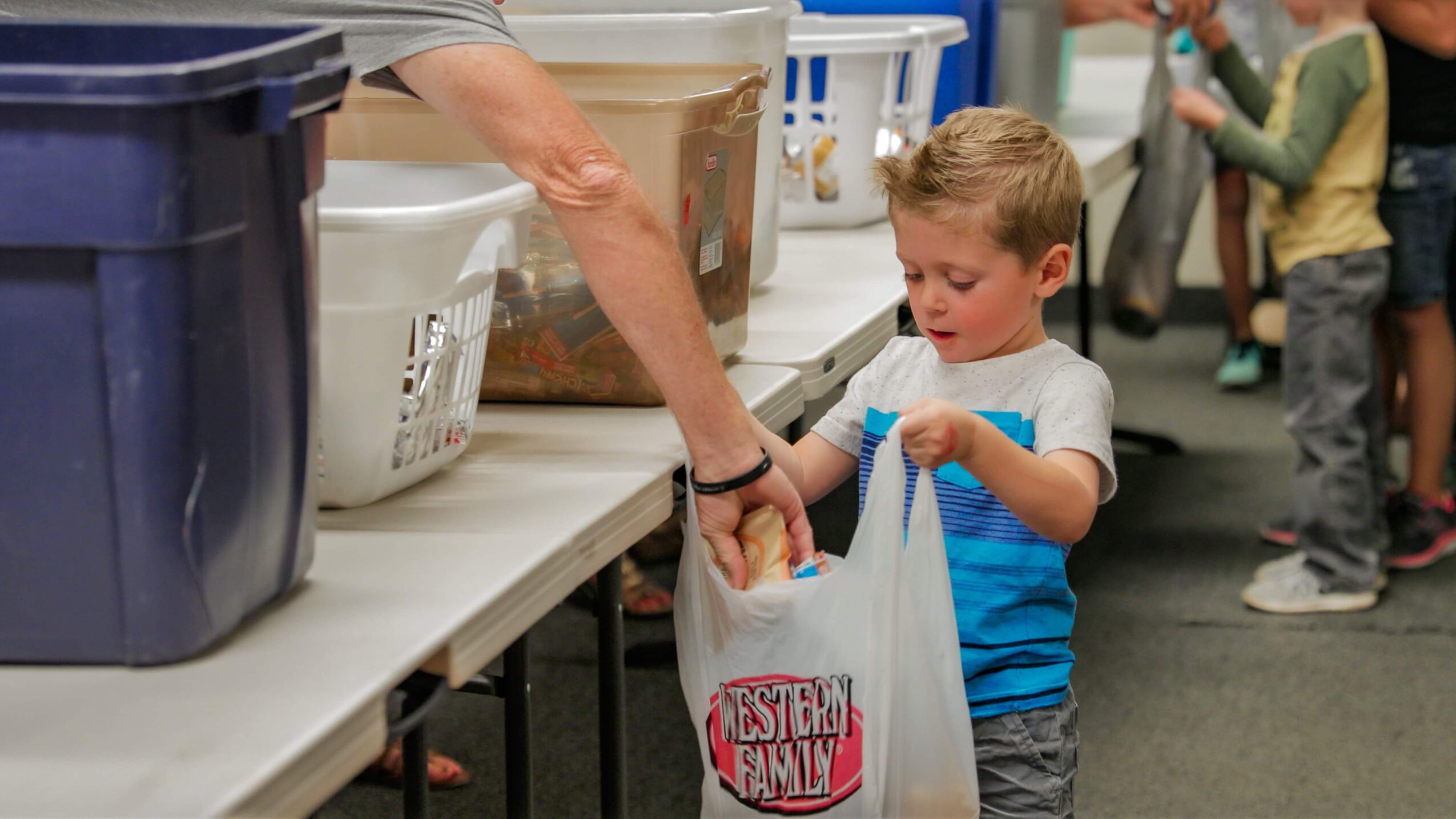 Child holding a bag at a donation table with an adult's hand nearby, in a busy indoor setting.