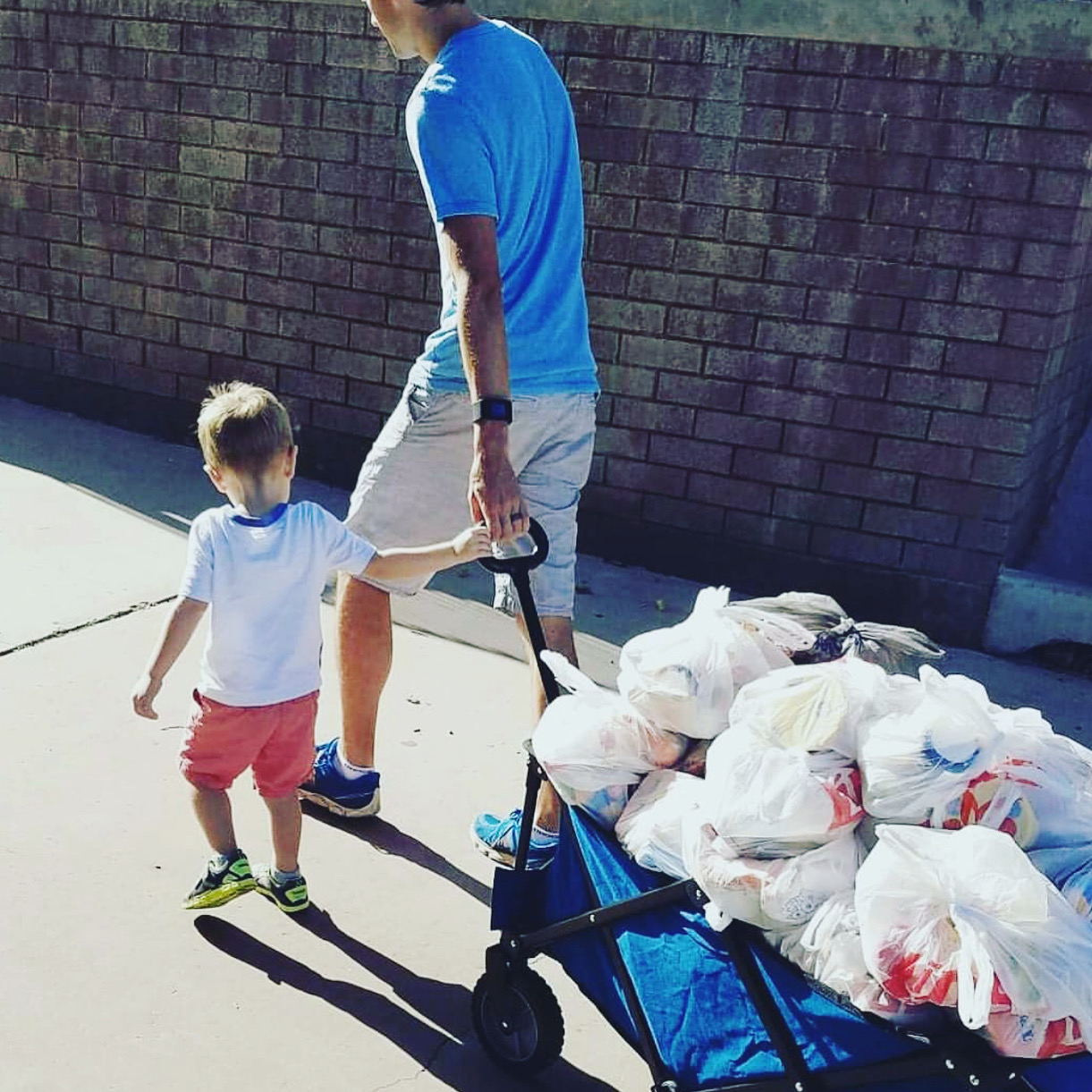 Adult and child walking with a cart filled with trash bags, outdoors near a brick wall.