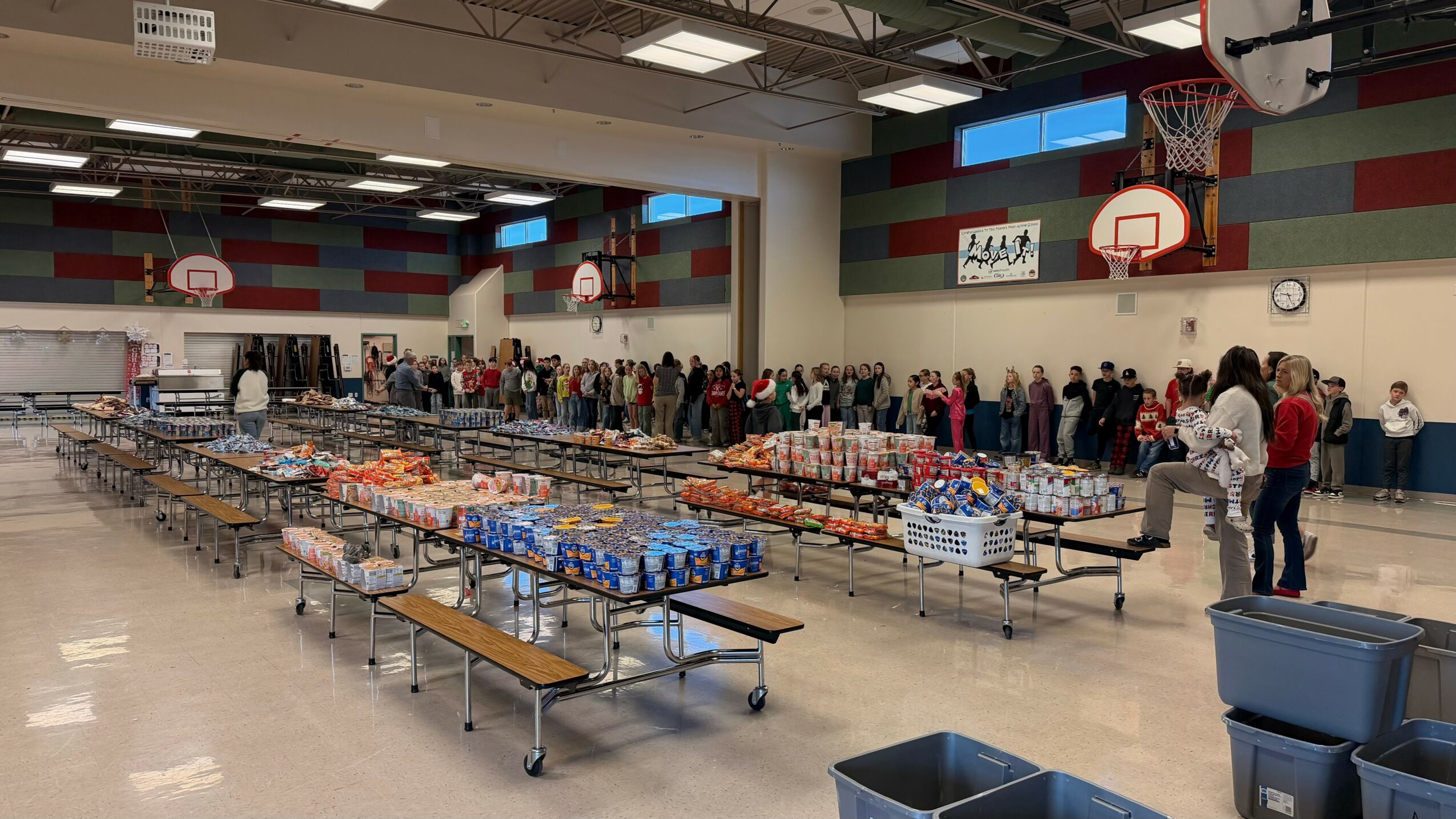Indoor gymnasium with tables of food and people in line, basketball hoops, and colorful wall panels.
