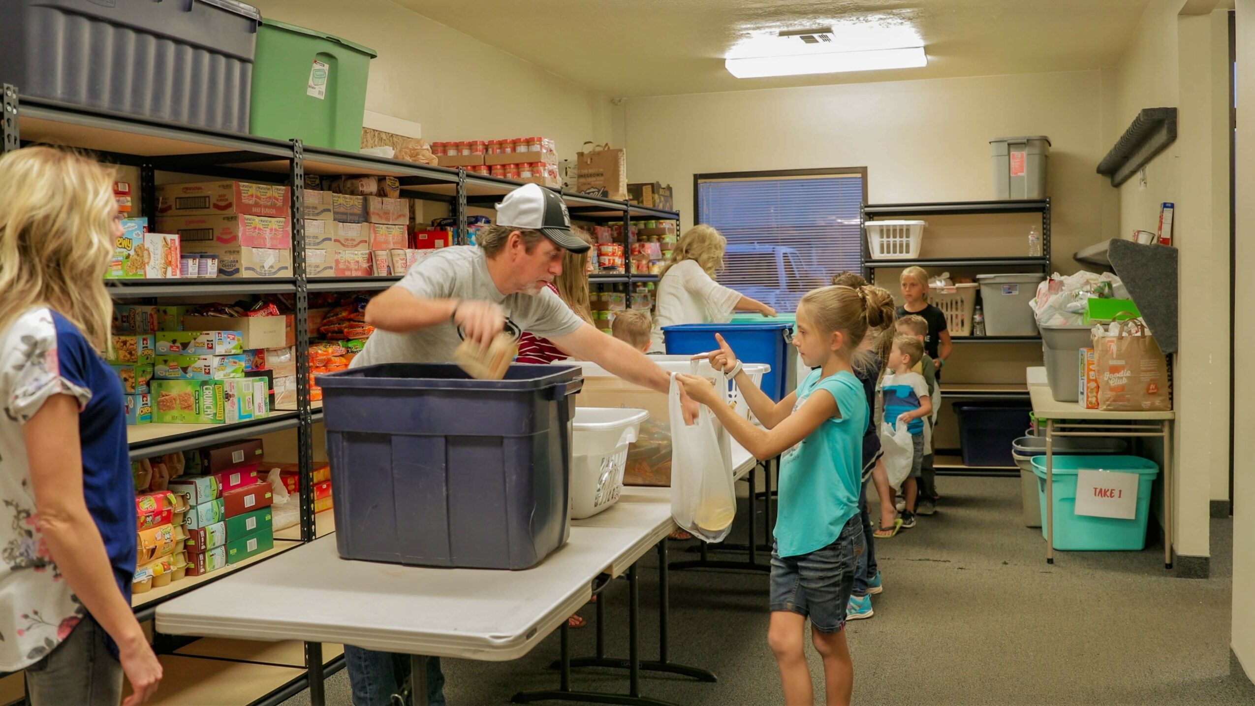 People distributing food to children in a room with shelves and boxes, some children reaching for food, adults assisting.