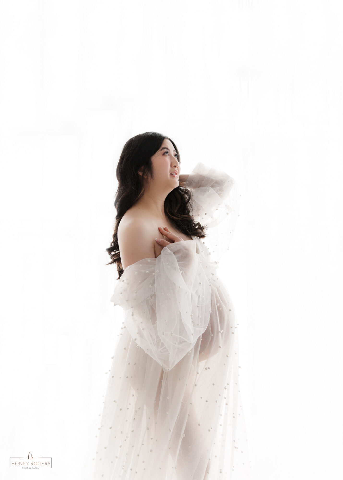 A woman with long dark hair in a white dress looks upward, holding her arms close to her chest, against a white background.