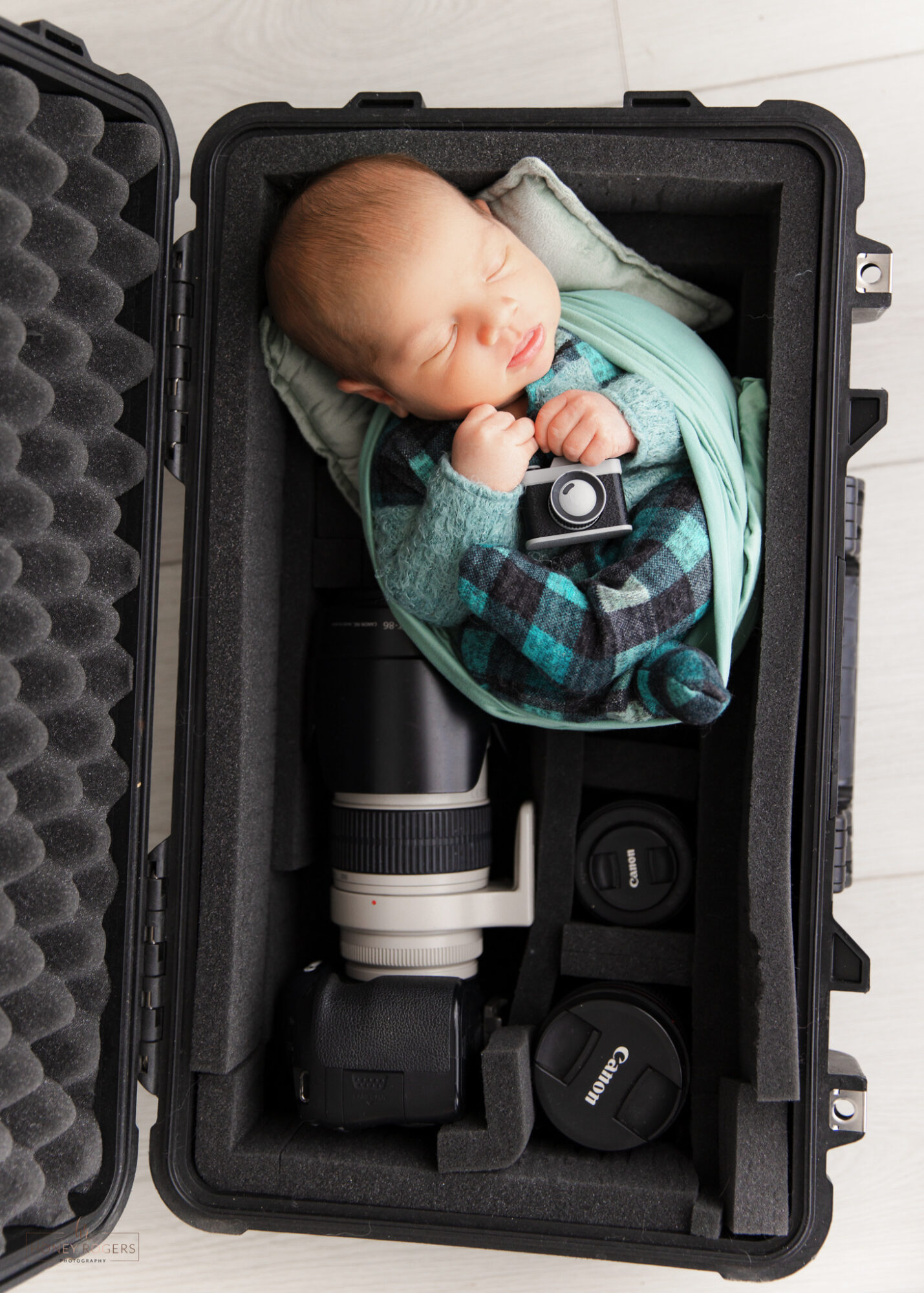Baby sleeping in a case with camera and lens accessories, wrapped in a blanket, on a white surface.